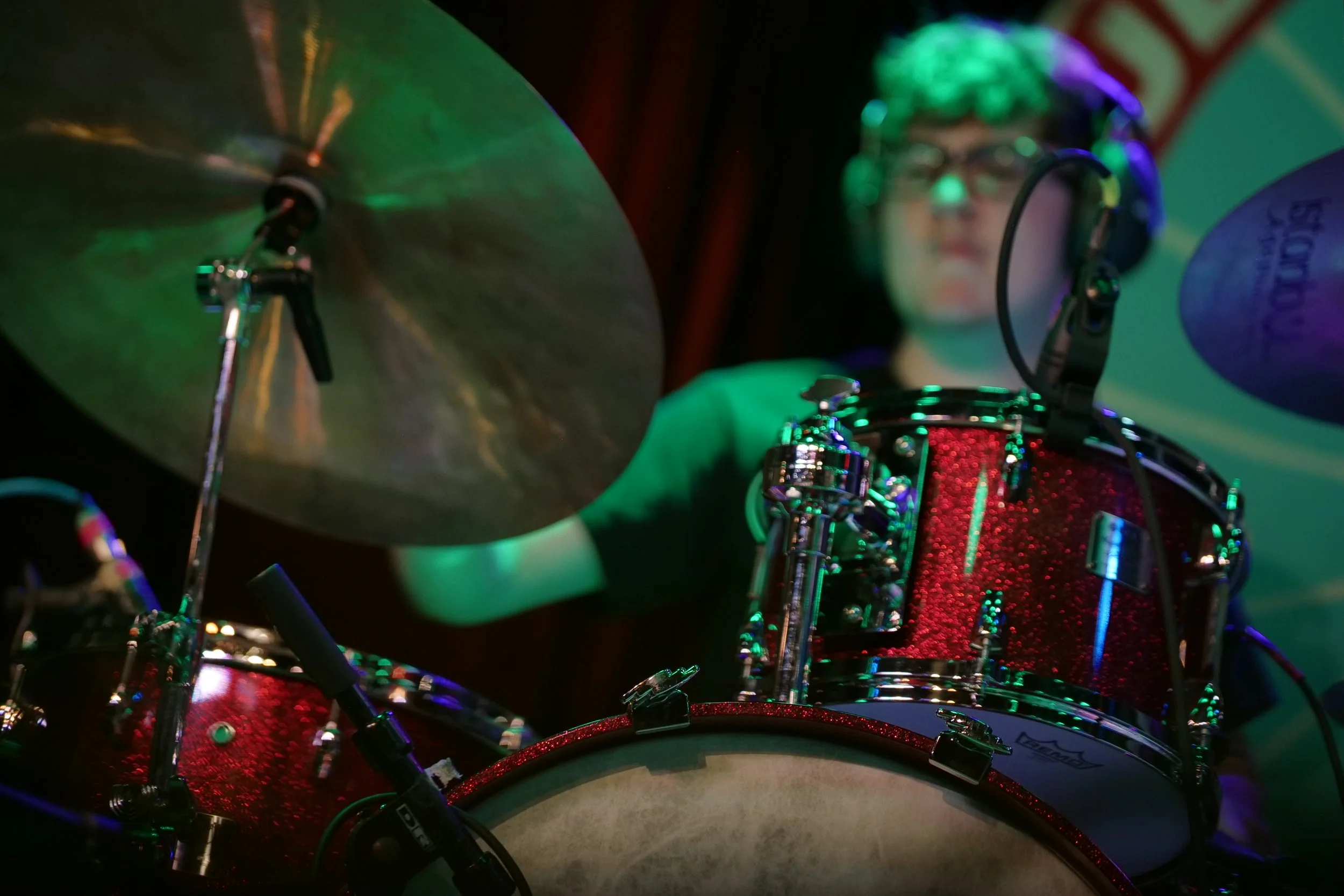 A young man playing drums, wearing glasses, with colorful stage lighting.