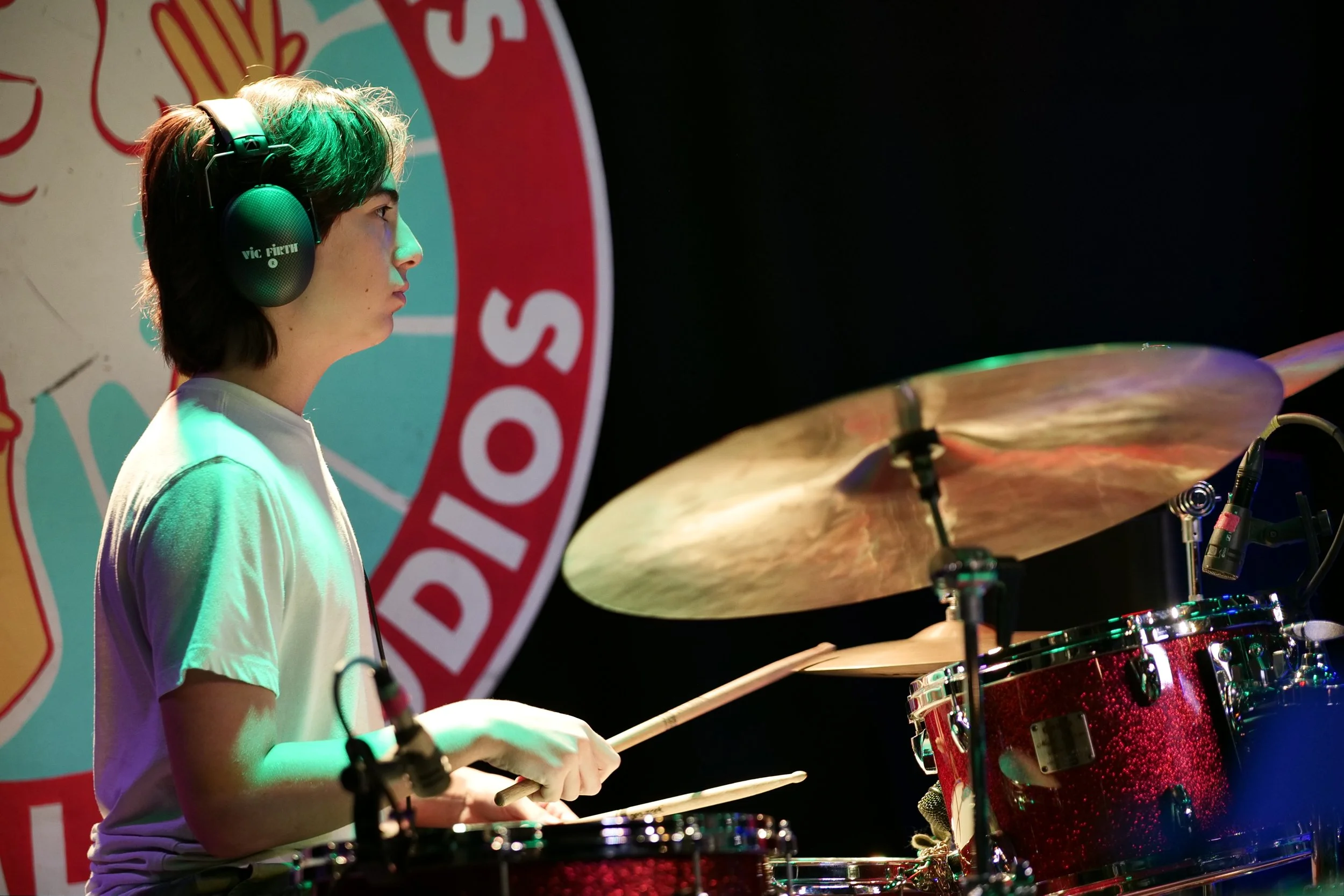 A young person playing drums on stage, wearing headphones, with a colorful background.