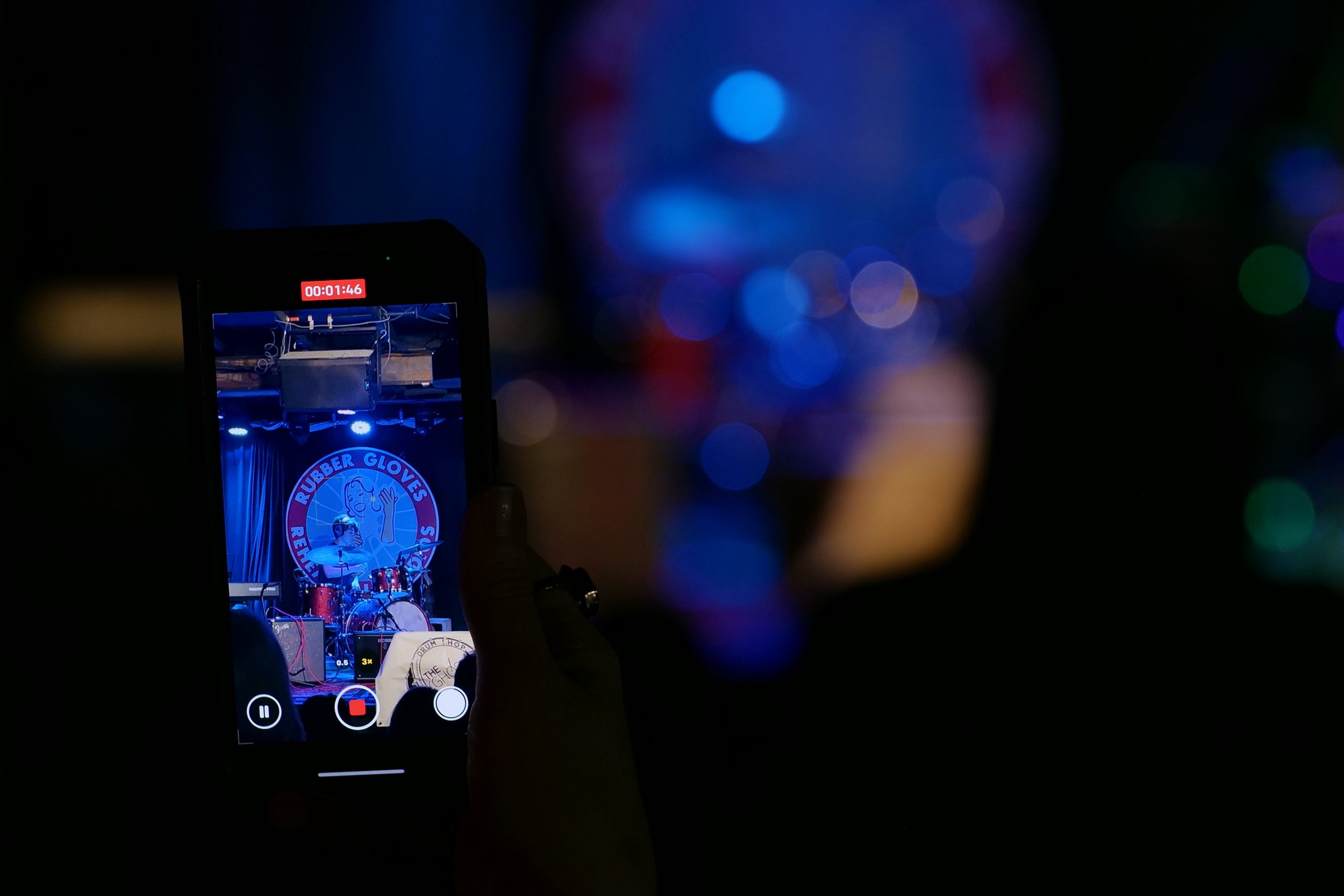 Person recording a live performance on a smartphone in a dimly lit venue with colorful blurred stage lights and a logo that reads 'Rubber Gloves Rehearsal Studio' on a backdrop.