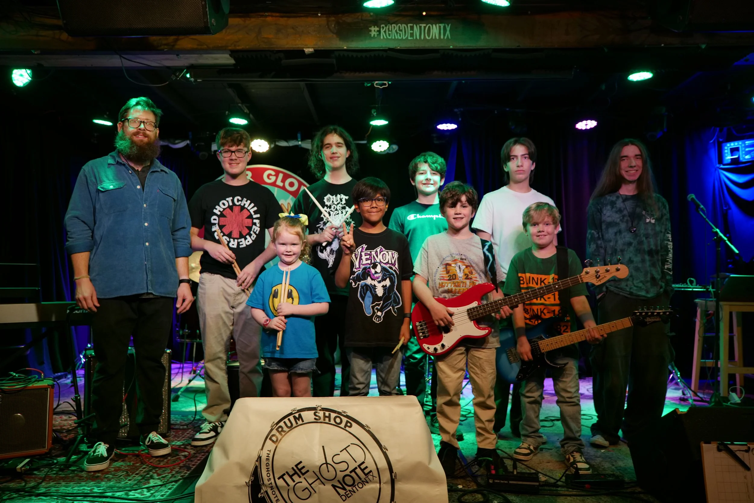 A group of eleven people on stage, including children and adults, posing with musical instruments and looking at the camera. They are under stage lights, with music equipment around them, and a drum shop banner in the foreground.