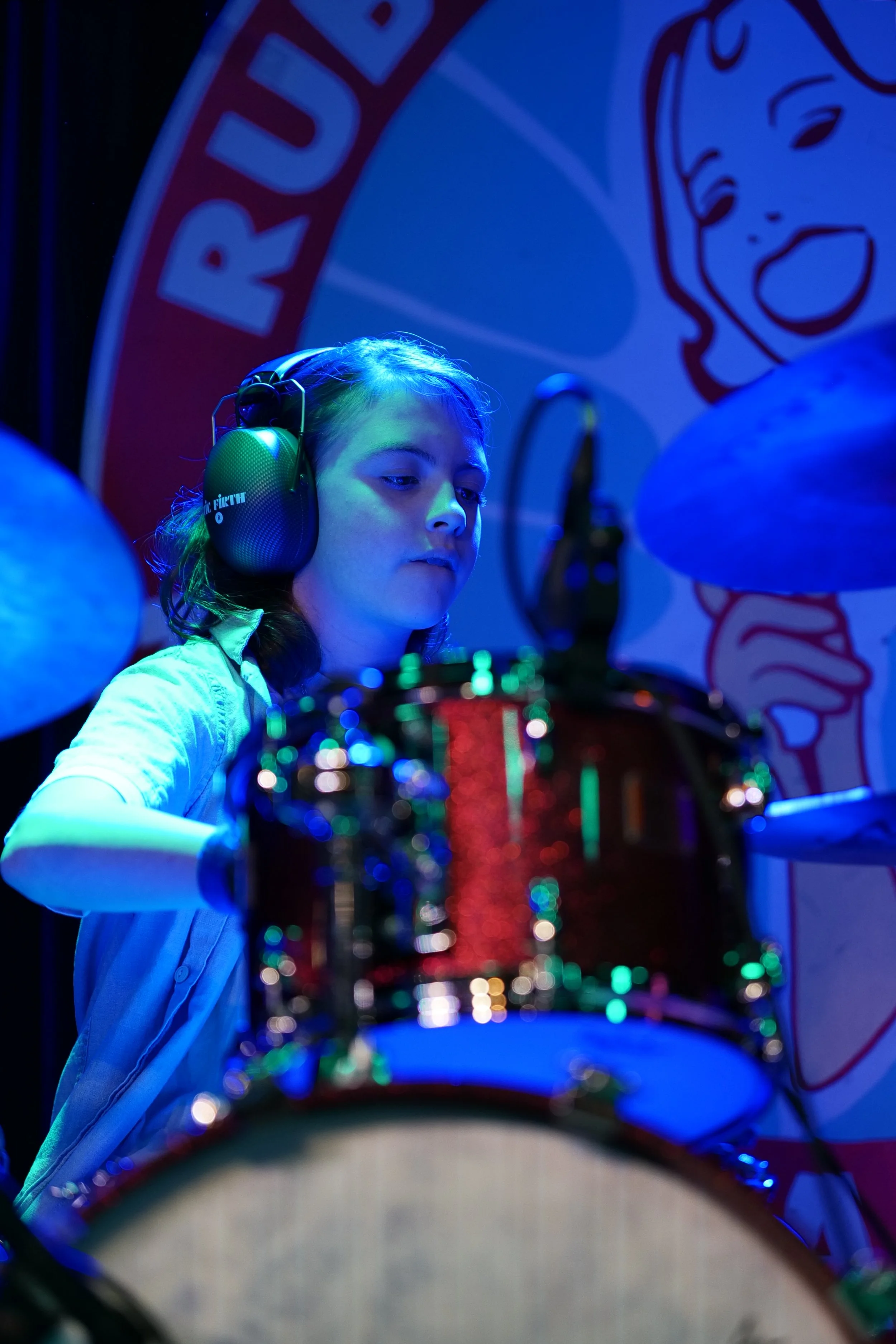 Young person with long hair wearing headphones playing drums on stage with a colorful background.