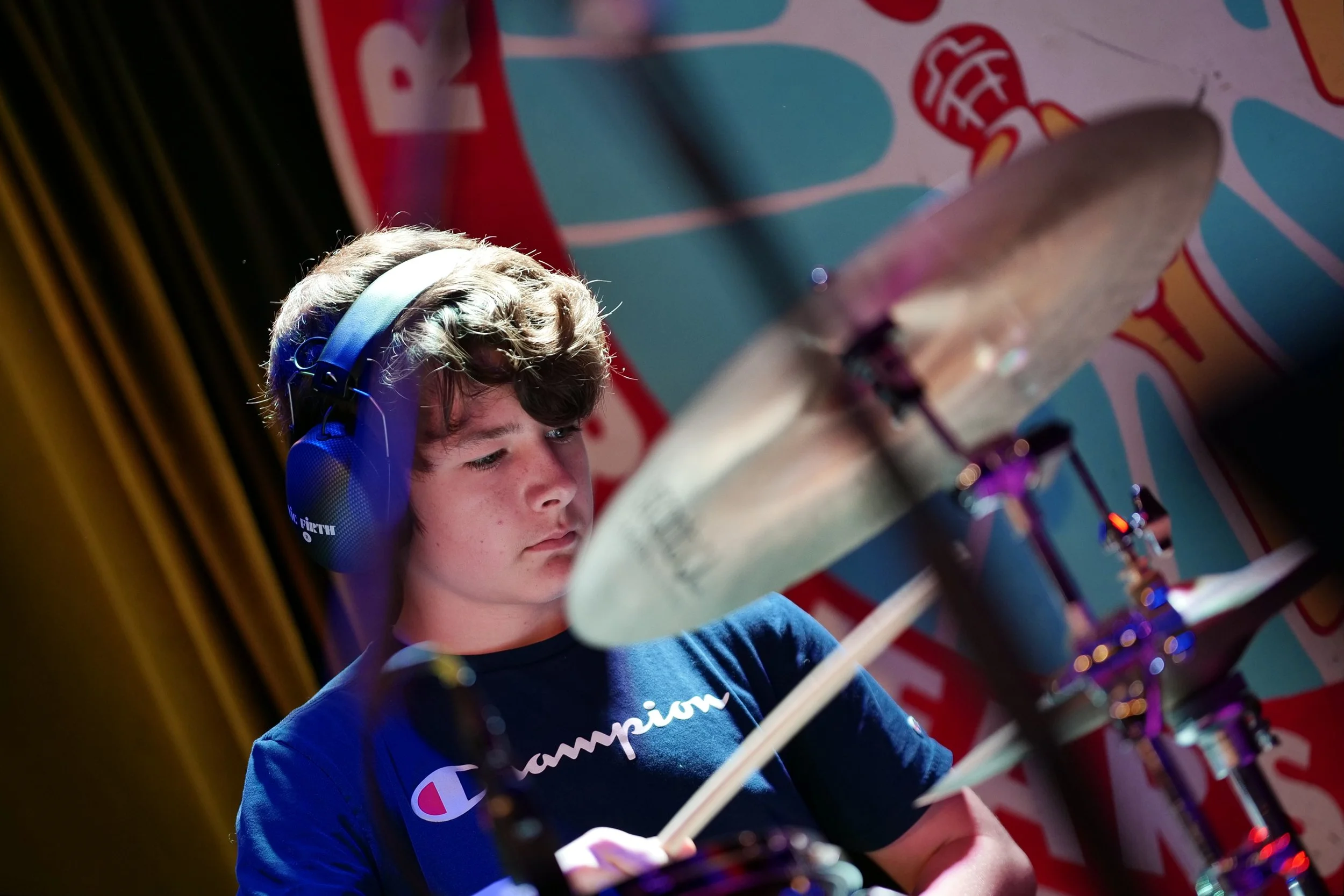 A teenage boy with curly brown hair wearing large headphones and a navy blue t-shirt with the Champion logo, focused on playing drums in a room with colorful wall art.