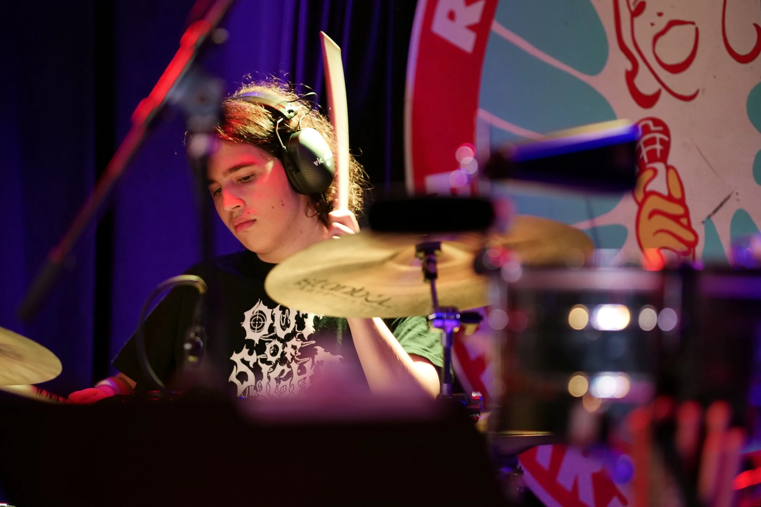 A young male drummer with headphones playing drums on stage, with a colorful backdrop and stage lighting.