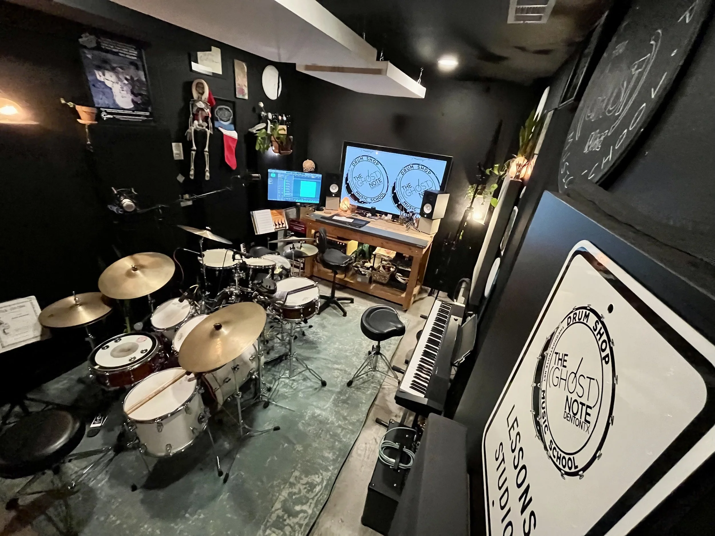 Music studio with electronic drum set, keyboard, two monitors displaying 'The Ghost Note,' mixing console, music stands, and wall decorations including a hanging skeleton and Christmas stocking