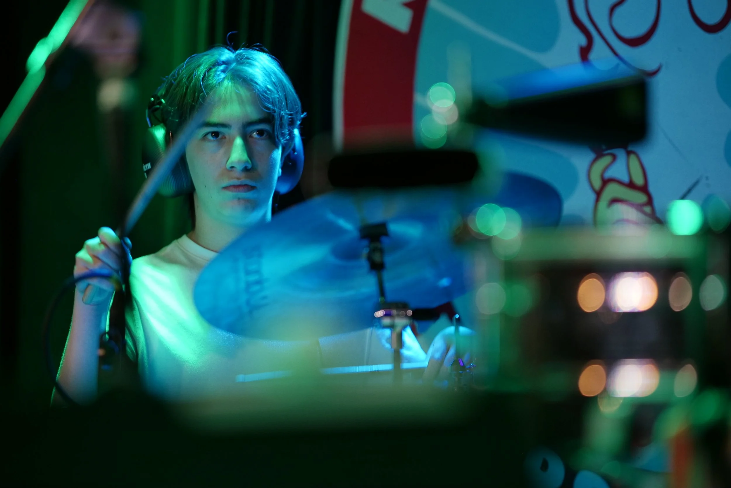 A young man with headphones playing drums in a colorful, dimly lit setting.