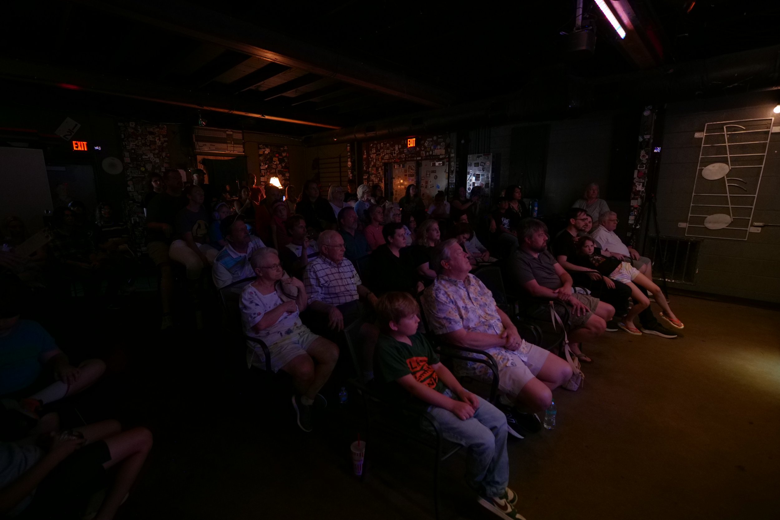 Audience watching a performance in a dark venue, seated in chairs facing a stage with musical note artwork on the wall.