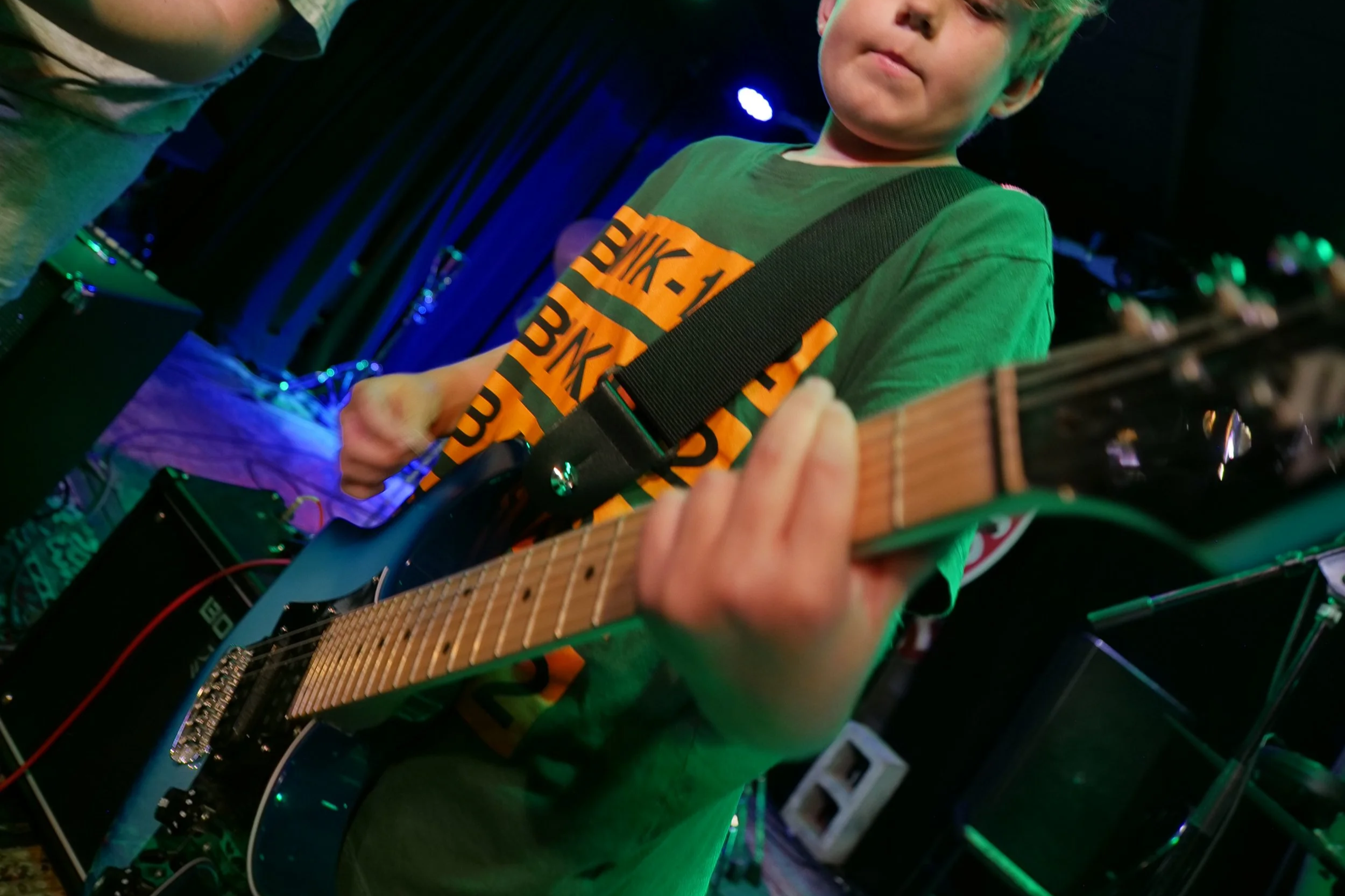 A young boy playing an electric guitar on stage in a dark room with blue and green lights. Guitar lesson Denton, TX