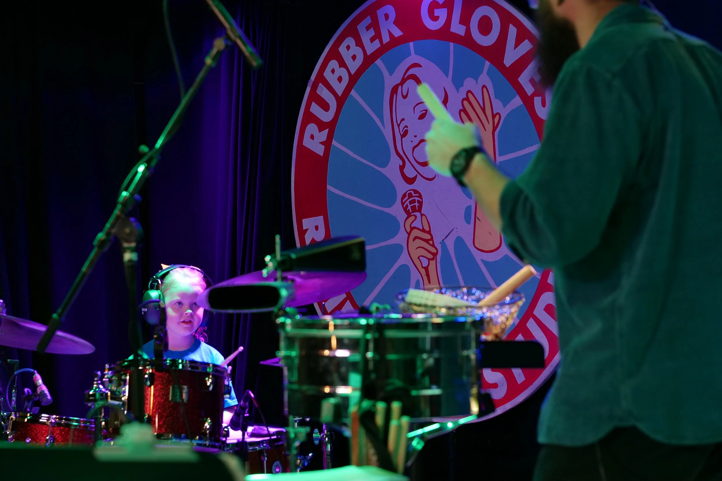 A young girl performing on a drum set during a live music event, with a large circular logo behind her featuring a cartoon person with a microphone, and the words 'Rubber Glove' in a semicircle. A man, partially visible, appears to be conducting or t