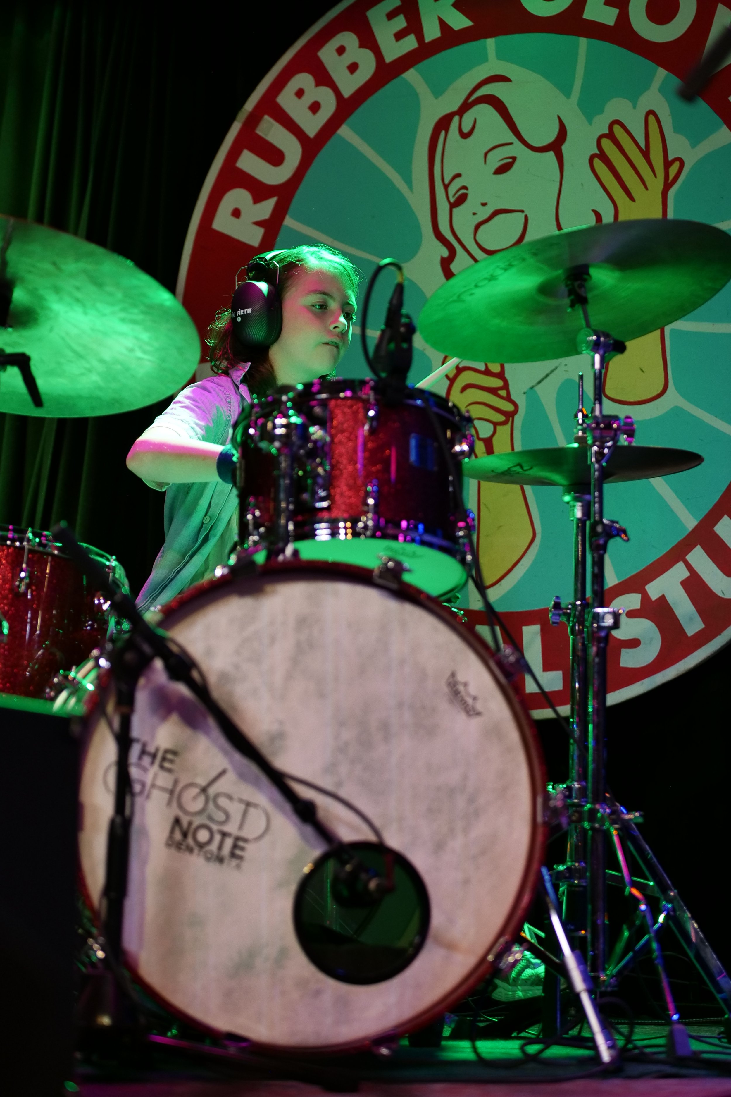A young girl playing a drum set on stage, wearing headphones, with a large colorful sign in the background that reads 'Rubber City' and features a smiling woman cartoon illustration.