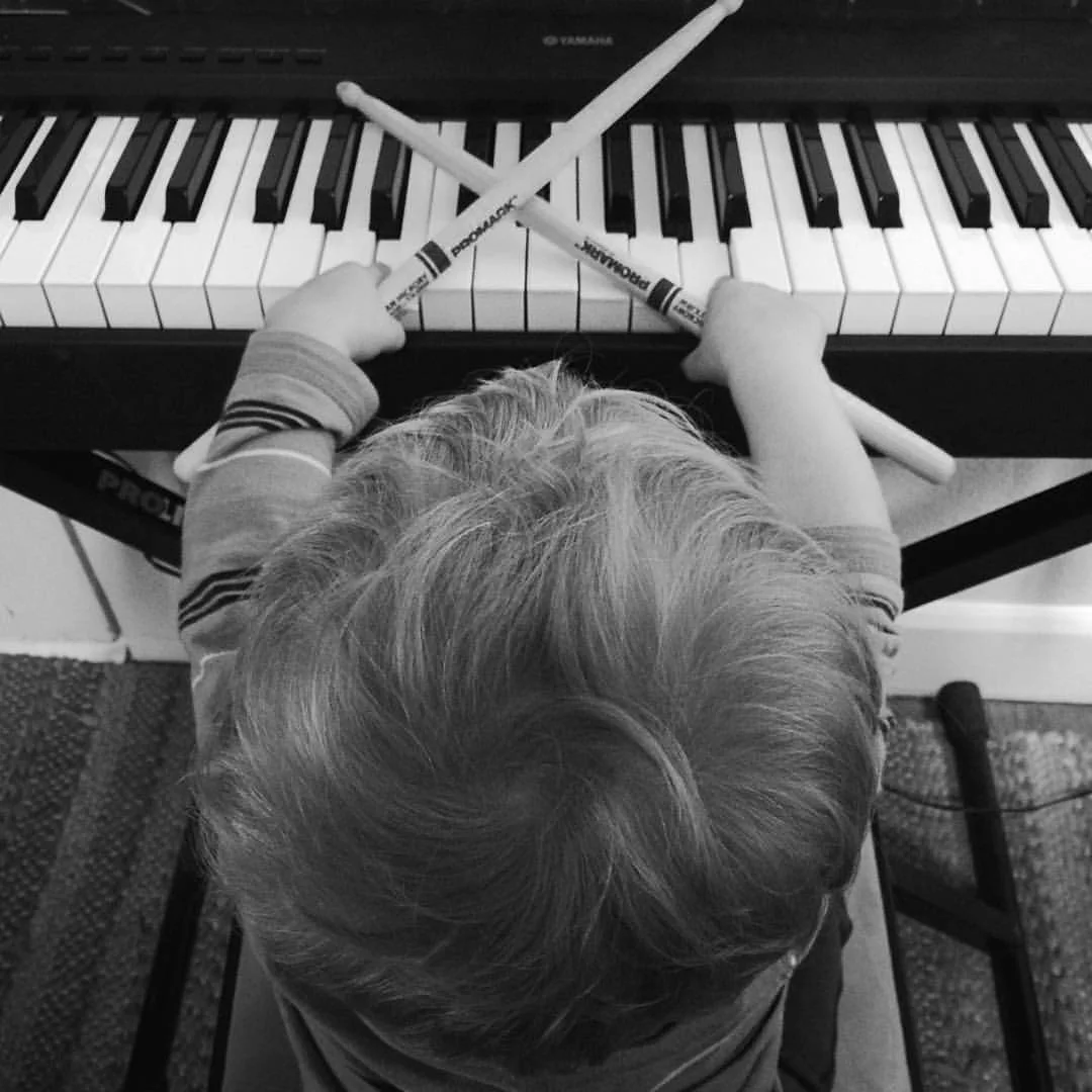 A young child with light-colored hair sitting at a piano, holding two large felt-tip markers crossed over each other, with the child's back facing the camera and the piano keys visible.