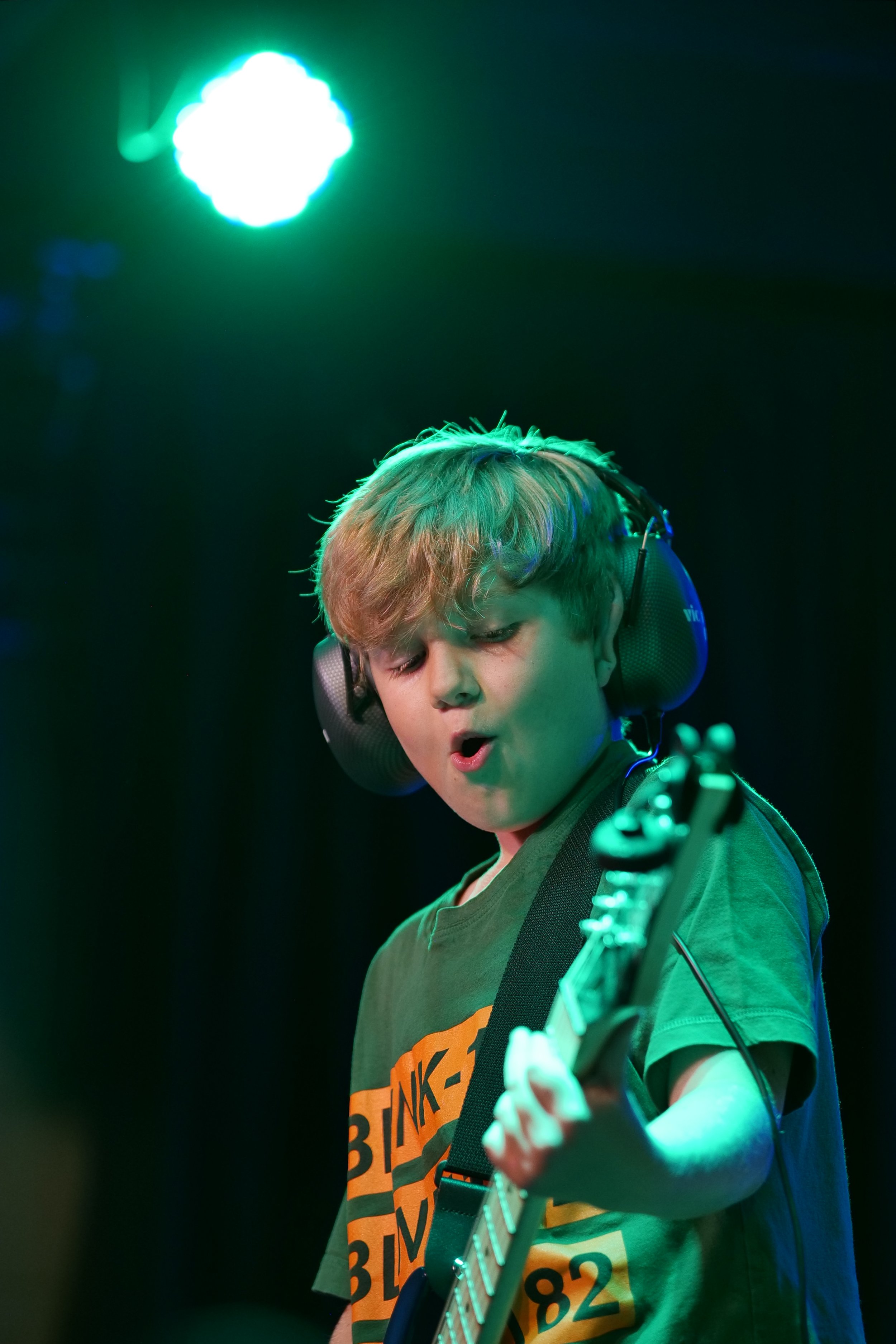 A young boy playing an electric guitar with ear protection, illuminated by colorful stage lights.