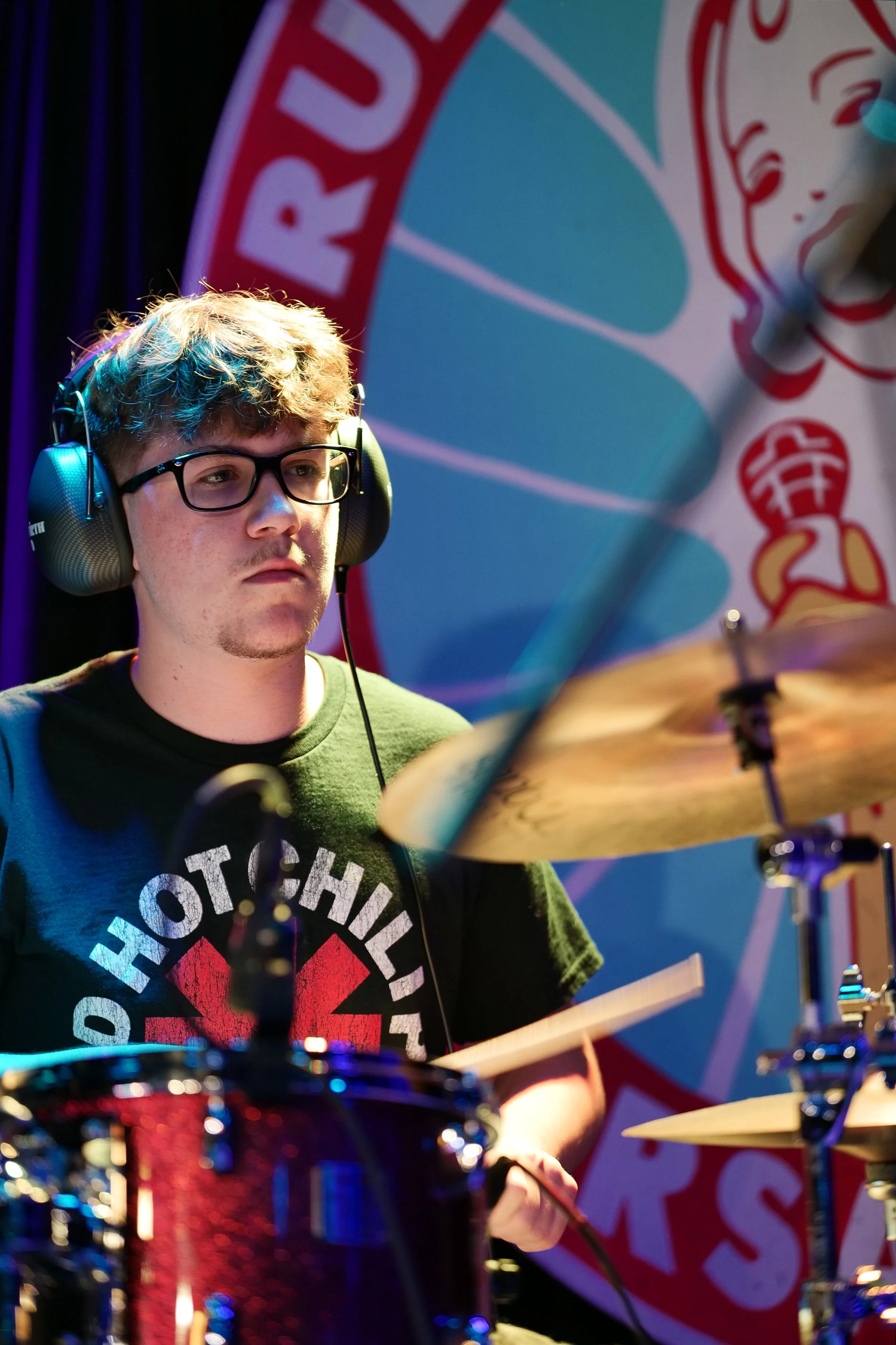 A young man with glasses and curly hair playing drums, wearing headphones and a Black Hot Chili Peppers T-shirt, against a colorful backdrop