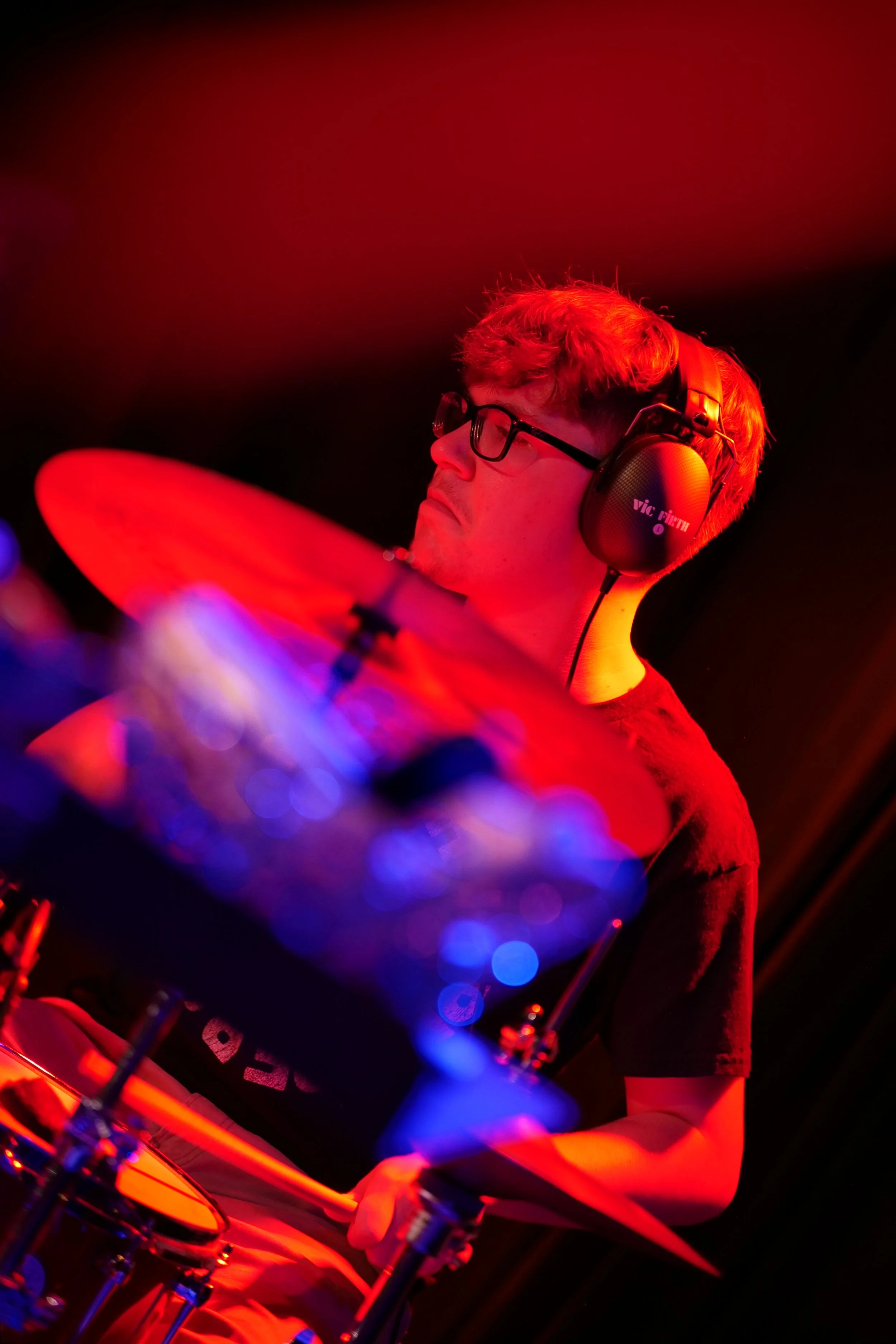 A young man with glasses and headphones playing drums in a dimly lit room with red and blue lights.