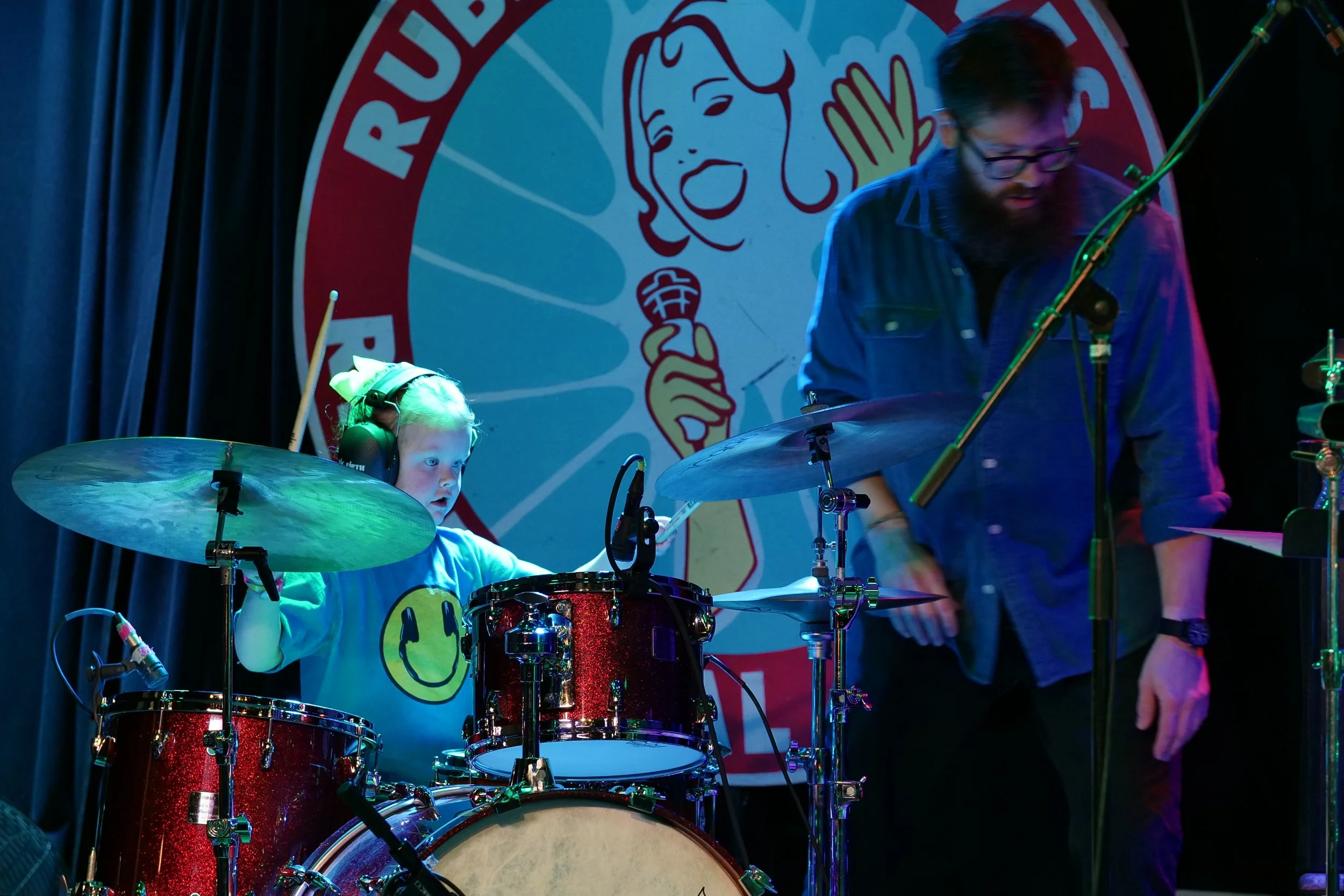 A young girl wearing headphones and a smiley face t-shirt playing a red drum set on stage, with a bearded man adjusting equipment nearby, in front of a poster with a cartoon face holding a microphone.