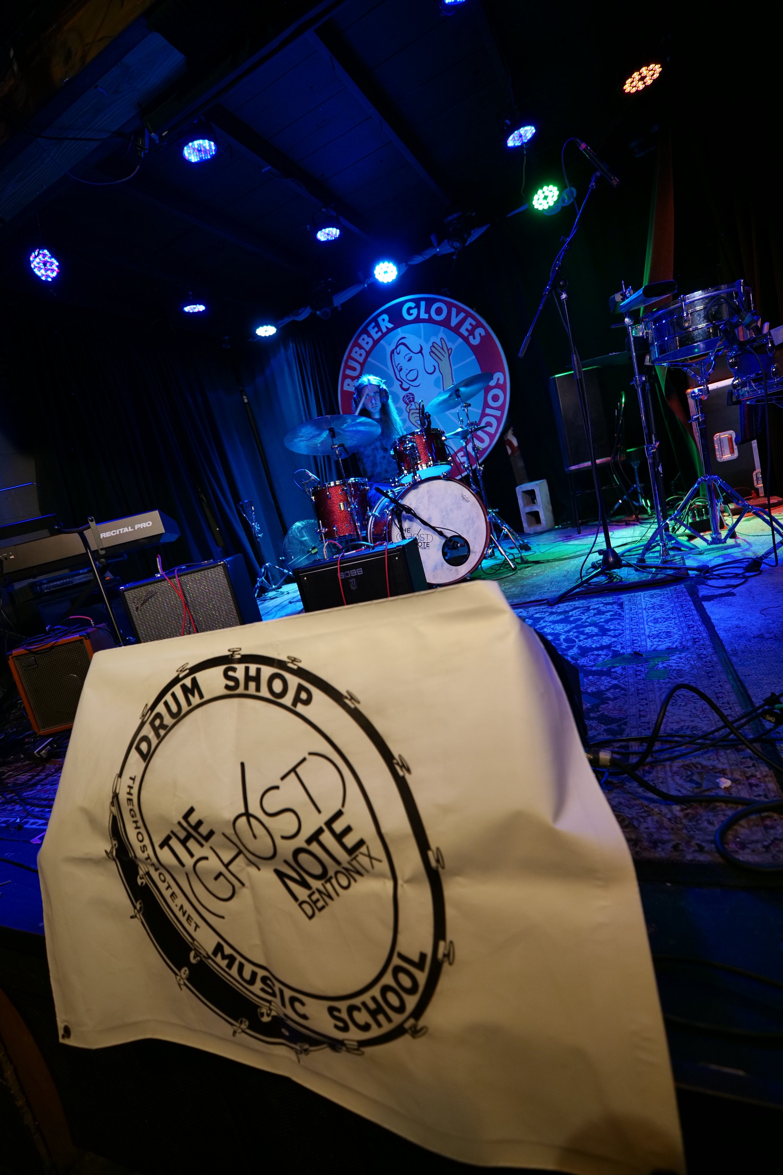 Stage with a red drum set, keyboard, and microphones, under blue and green lights, in front of a black curtain and a circular sign that reads 'Rubber Glove's Side Show'. A paper with 'The Ghost Note' and 'Music School' is in the foreground.