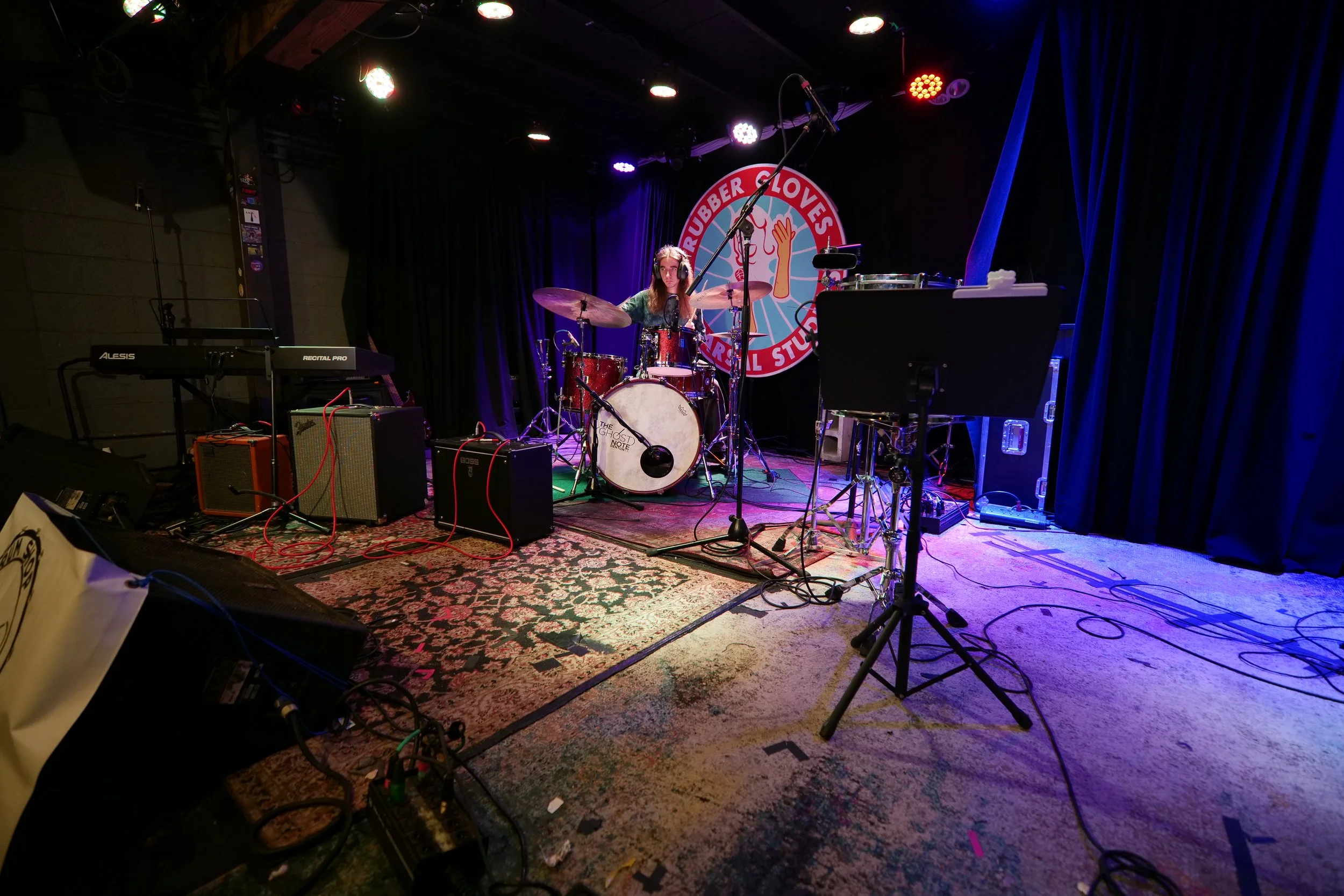A person plays drums on a stage with a sign reading 'Rubber Gloves Cultural Studio' in the background. The stage is set with a keyboard, amplifiers, and various musical equipment, with colorful stage lights overhead.