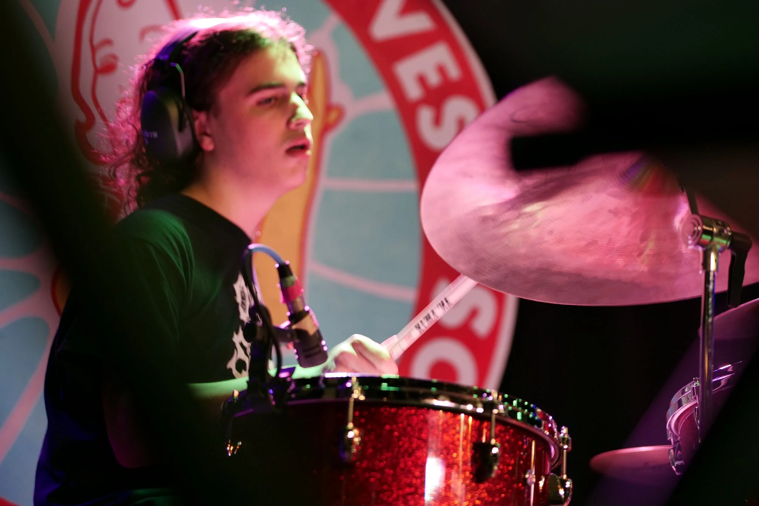 Young woman playing drums with headphones on, focused on her performance, in front of a colorful background with a circular logo.