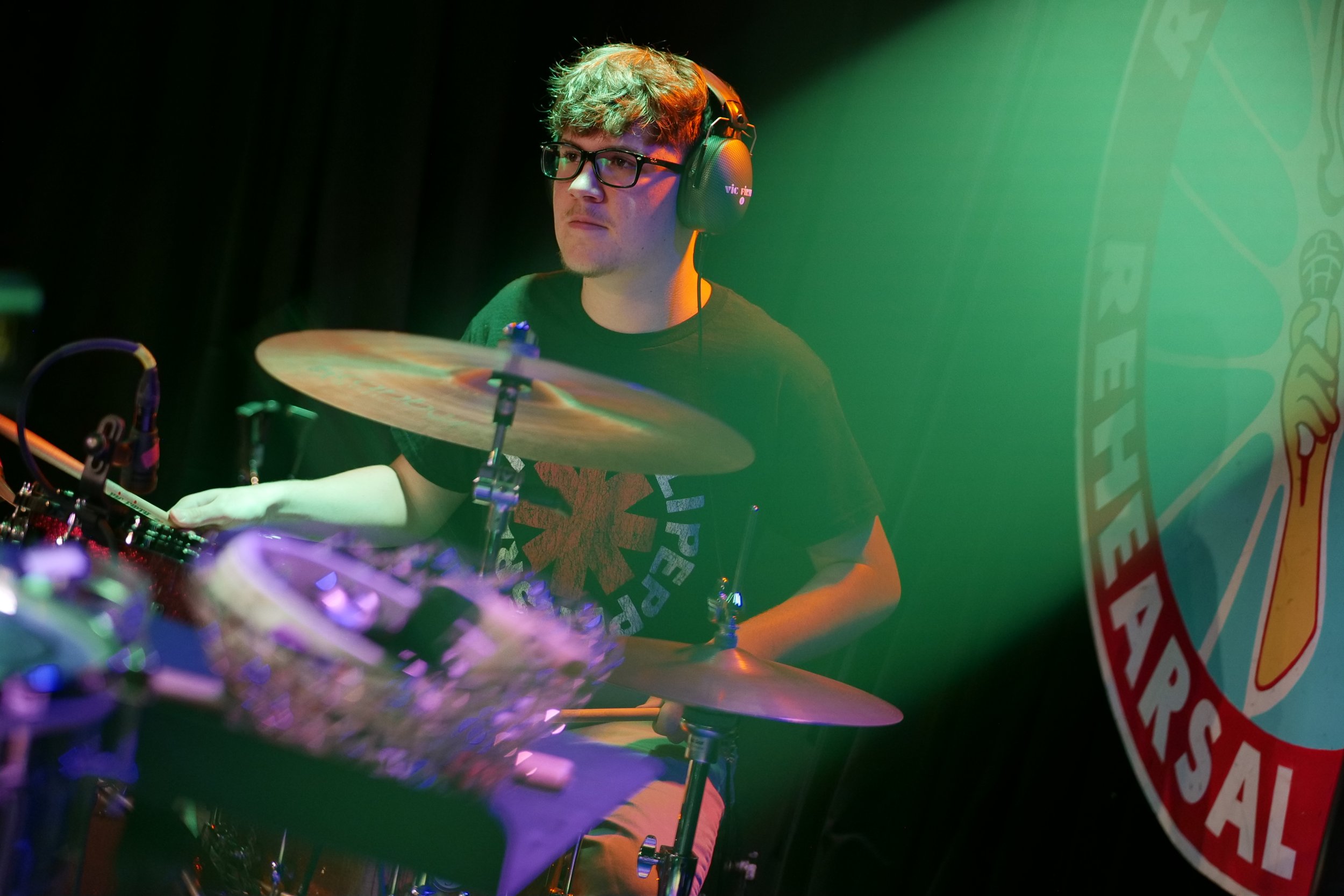 A young man with glasses and curly hair playing the drums on stage, wearing headphones, with a large circular logo on the right side of the image.