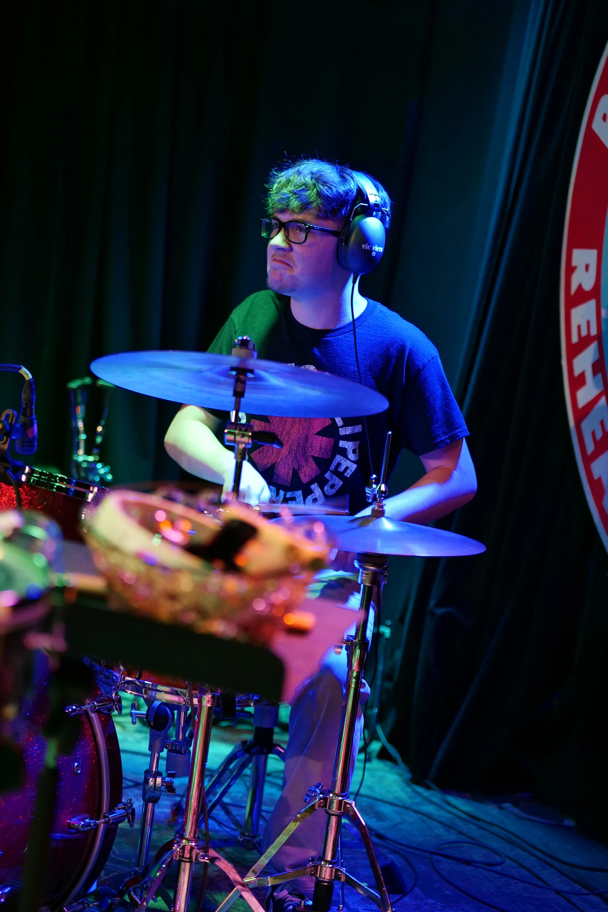 A young man wearing headphones and glasses, playing drums on a stage with colorful lighting.