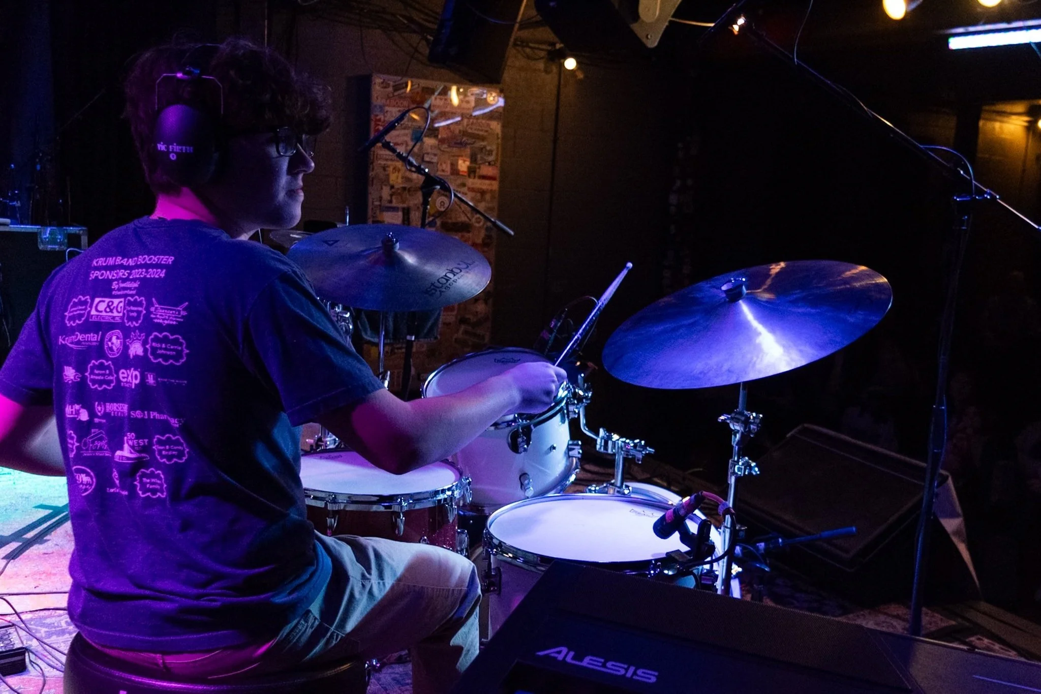 A young man with curly hair wearing glasses and headphones playing a drum set on stage in a dimly lit venue.