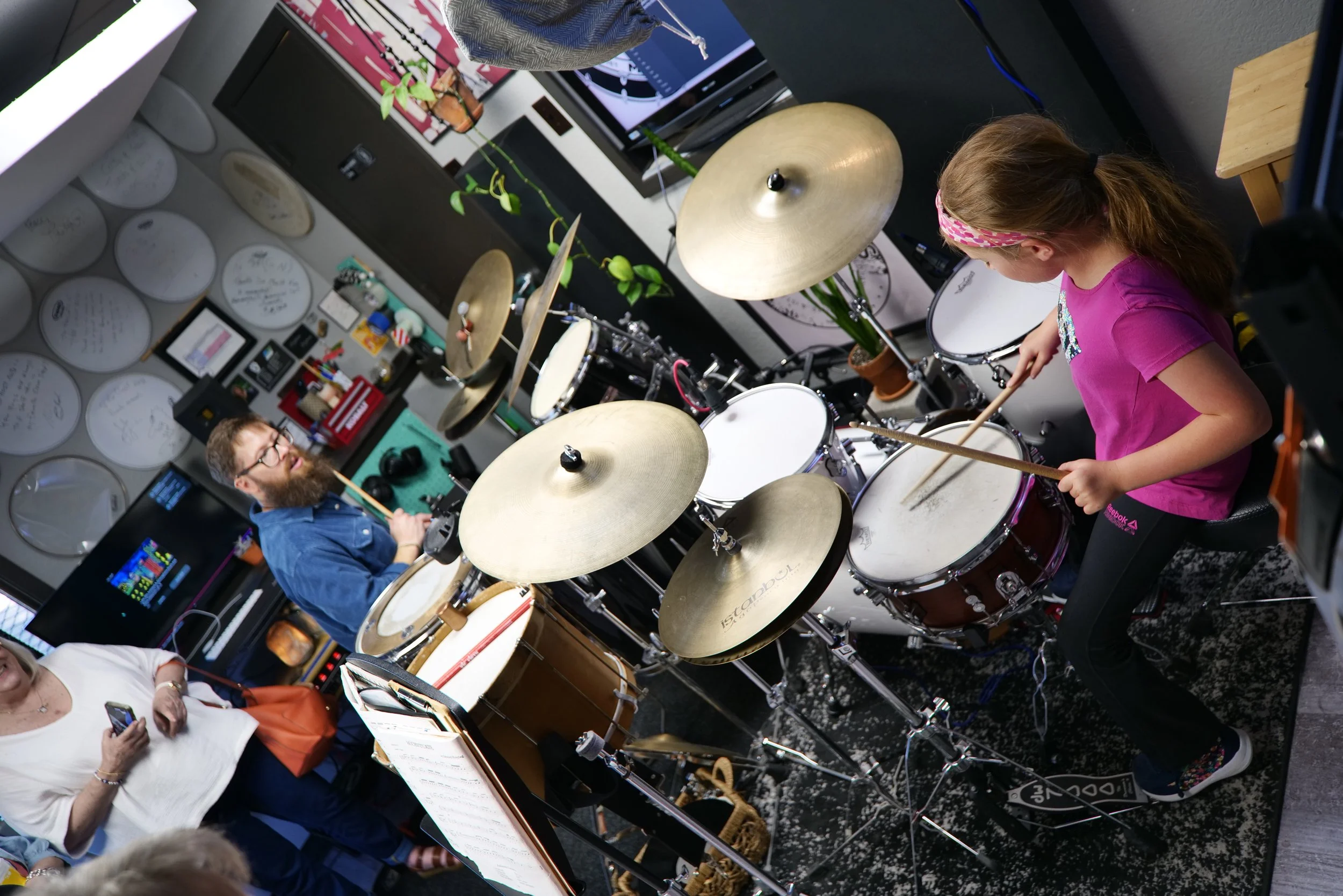 A girl in a pink shirt and black pants playing an electric drum set with a man in glasses and a beard watching. The room has wall decorations with white plates, a TV, and various items on shelves.
