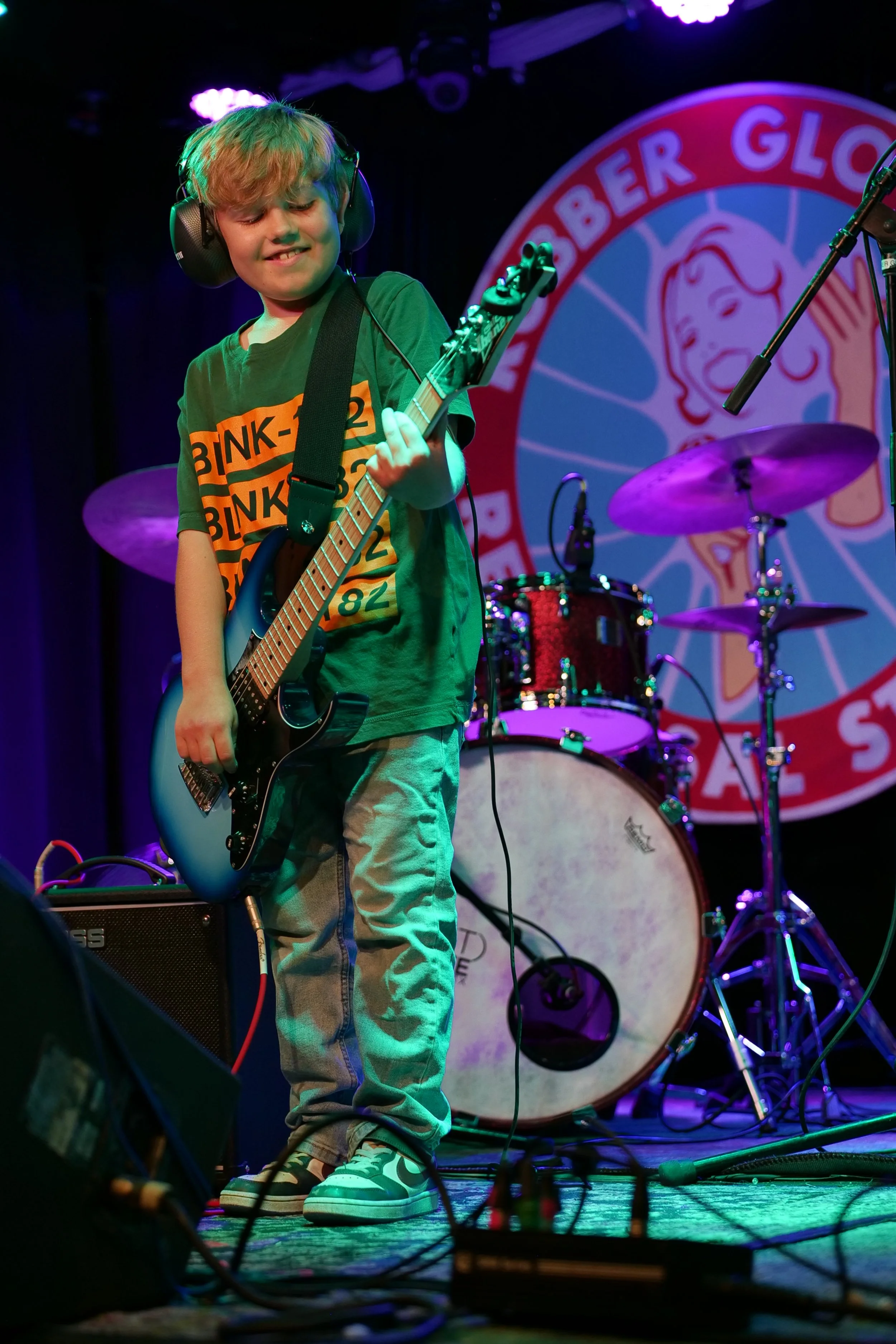 A young boy playing an electric guitar on stage during a live performance, with a drum set and a colorful background banner behind him.