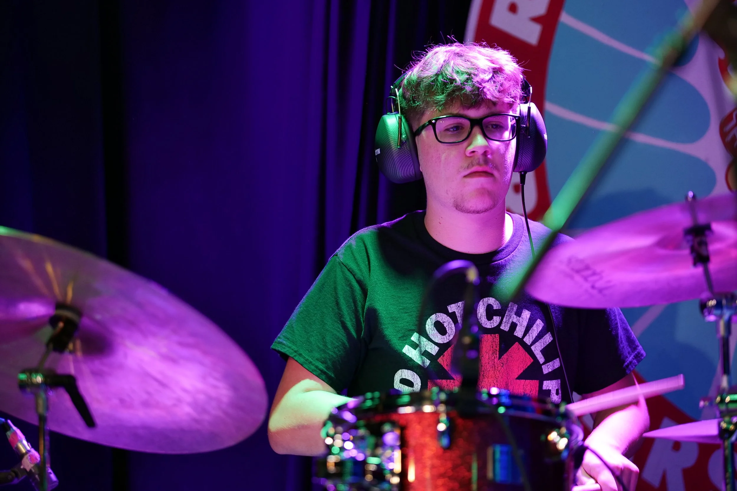 Young man playing drums on stage, wearing headphones and glasses, with a logo T-shirt, under purple and green stage lights.