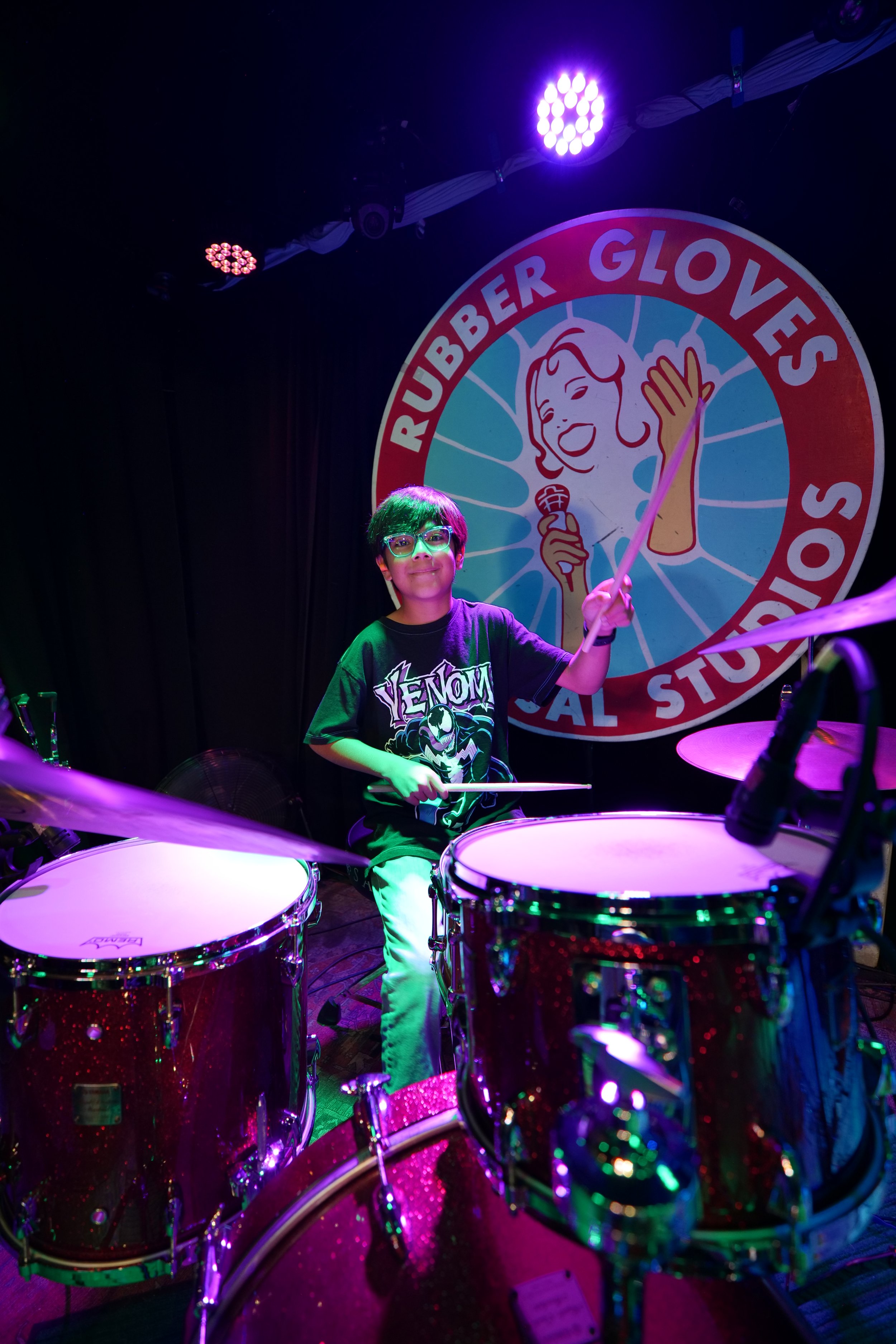 A young boy with glasses wearing a Venom t-shirt playing drums on stage at Rubber Gloves Laboratory. Office, stage lights, and a circular sign with a retro-style illustration of a woman with a microphone in the background.