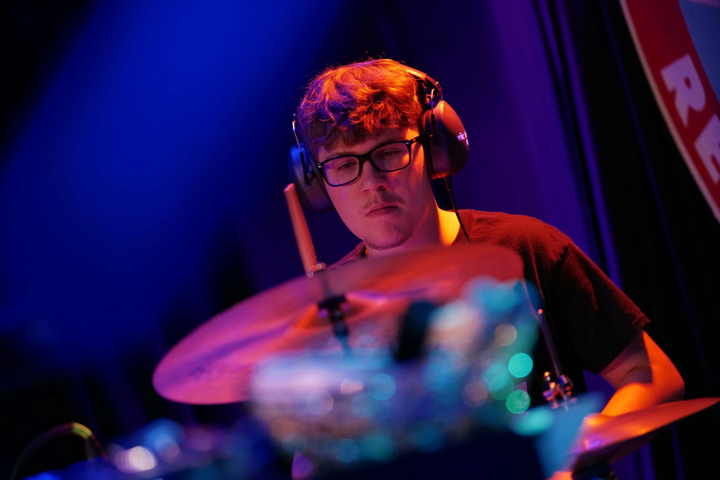 A young man with glasses and headphones playing drums on stage under colorful lighting.