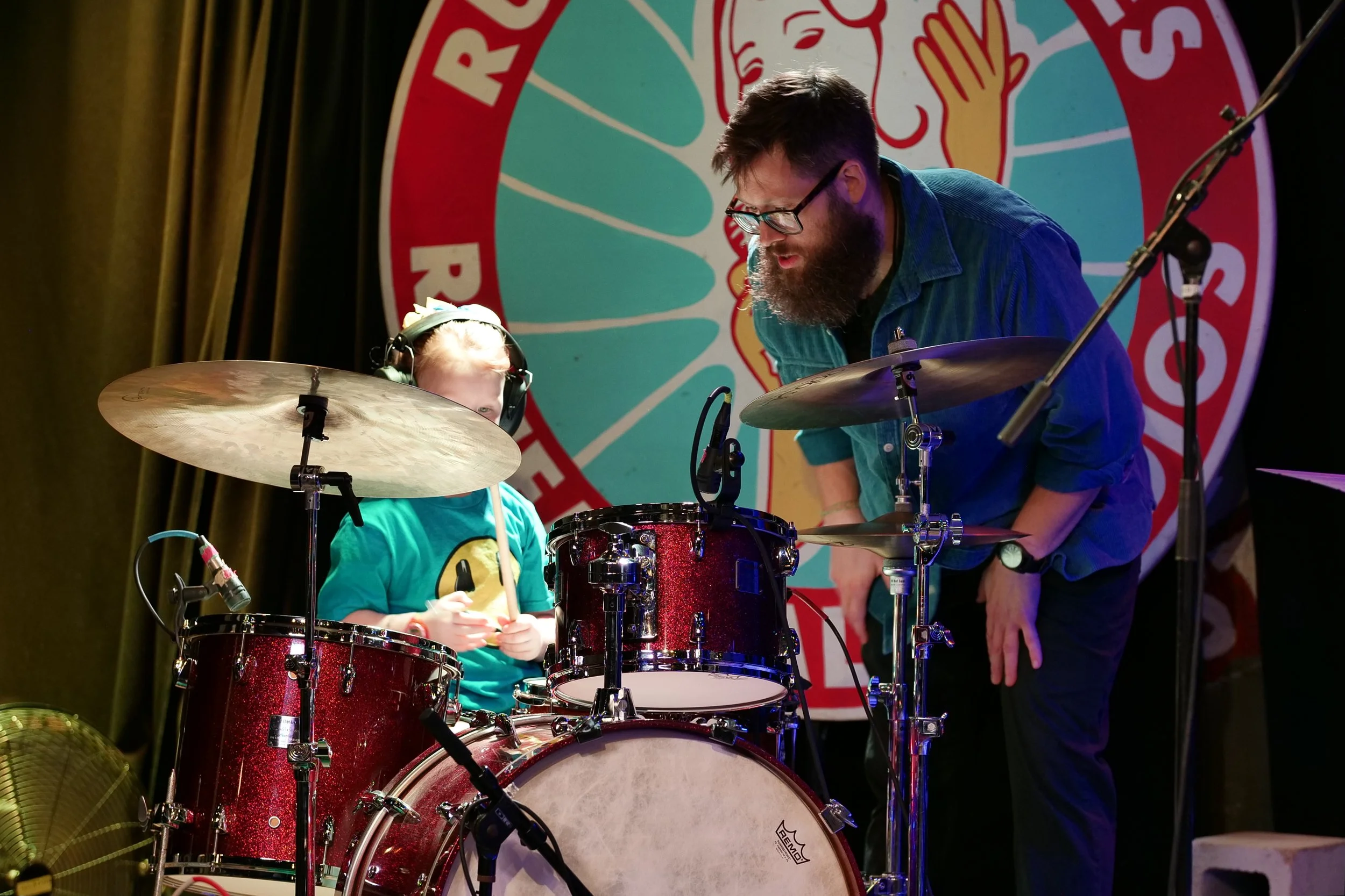 A young girl and a man are behind a red drum set on a stage, with a large circular logo in the background. The girl wears headphones and a teal shirt, while the man leans over the drums, talking to her. The stage has dark curtains and stage equipment