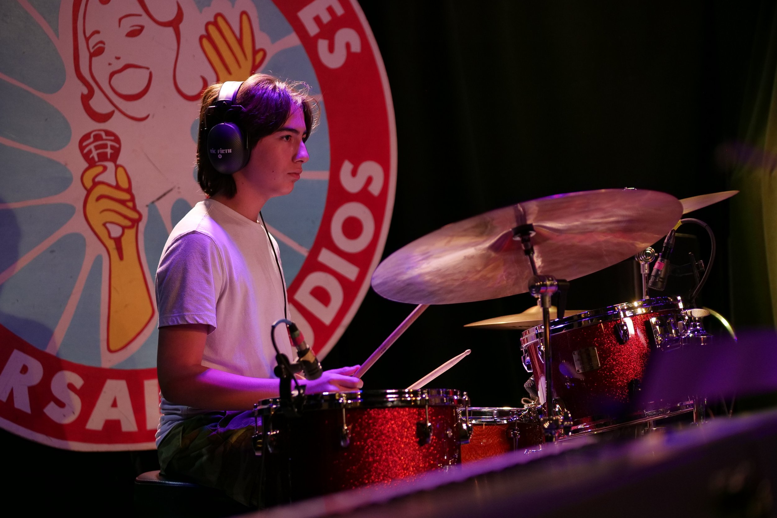 A young man with shoulder-length dark hair, wearing a white t-shirt and headphones, playing a red drum set on stage. Behind him is a large sign with a cartoon woman holding a screwdriver and the words 'Studio SOS' in a circle.