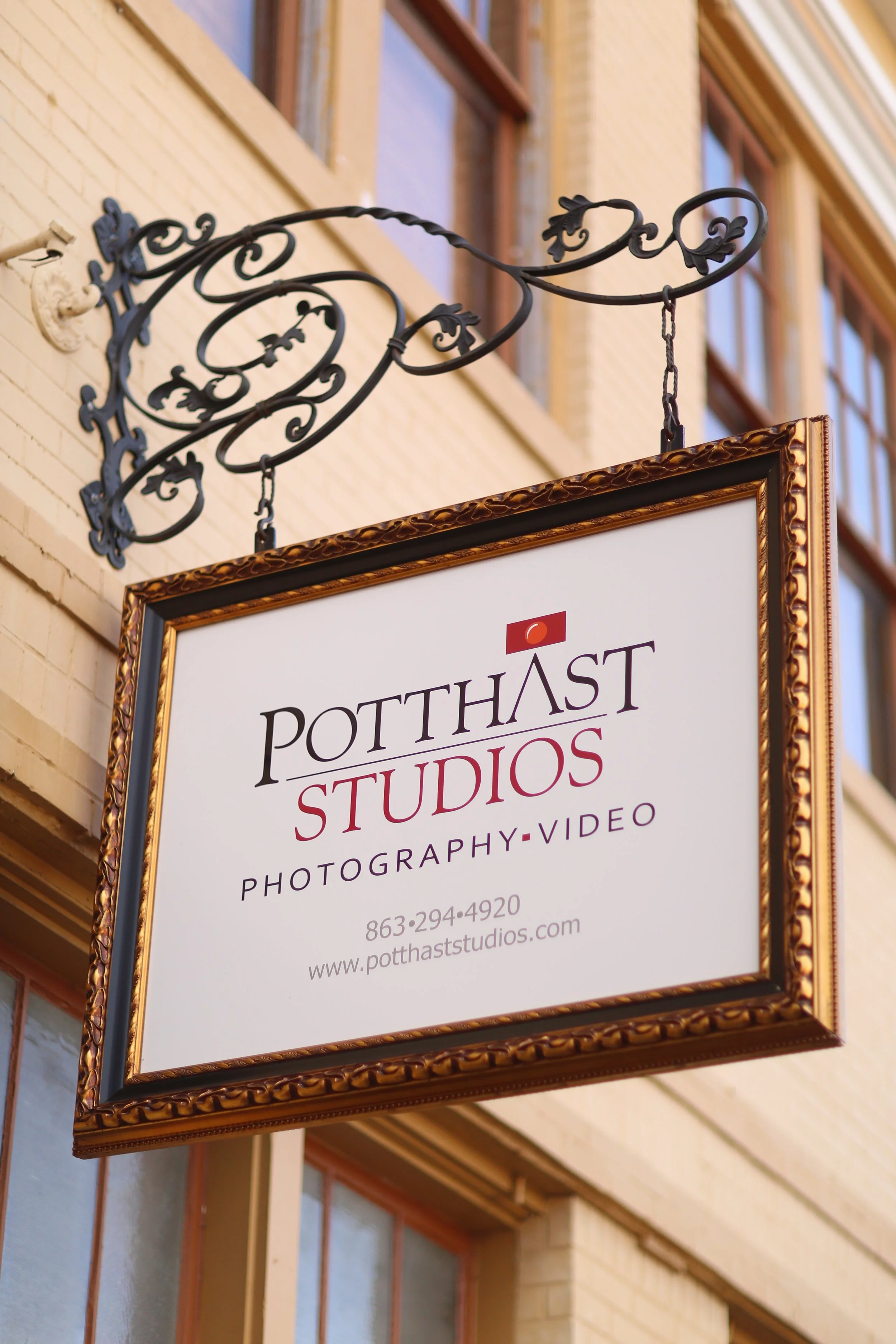 Business sign hanging from a decorative wrought iron bracket on the exterior of a brick building, advertising Potthast Studios for photography and video services.