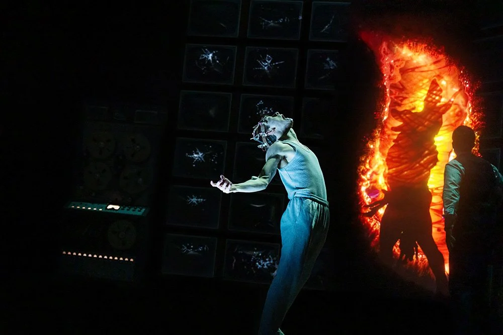 A young man, wearing a metal device on his head, looks up in pain. Behind him, there is a vortex of fire and an older man is looking into the flames.