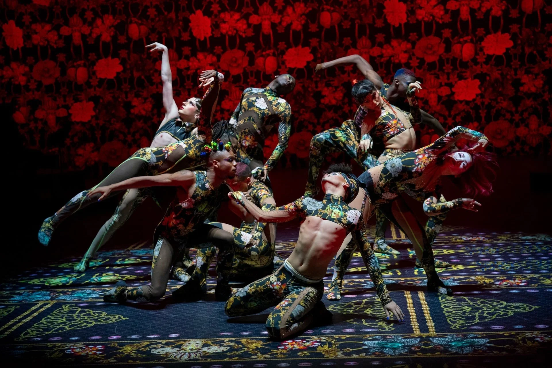 Dancers in various positions, dressed in floral clothes, dancing in front of a red floral screen.