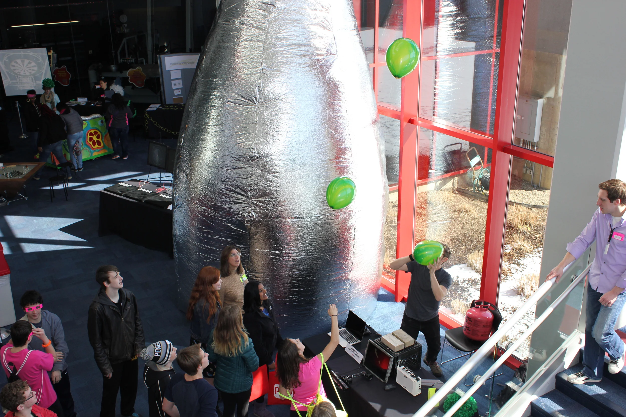 Students interact with an installation by artist Dan Wilcox pictured holding a green ballon. Wilcox uses an Xbox Kinnect to detect where balloons are in relation to the camera. The movement and proximity of the balloons affects different sound outputs. Wilcox's wife, Anika, has installed one her large mylar inflatables pictured centered.