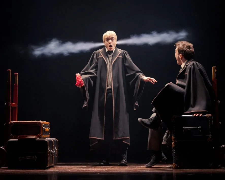 A young blonde teenager, wearing black robes, reacts to smoke coming out of his ears. In his hand, he is holding a bag of candy.