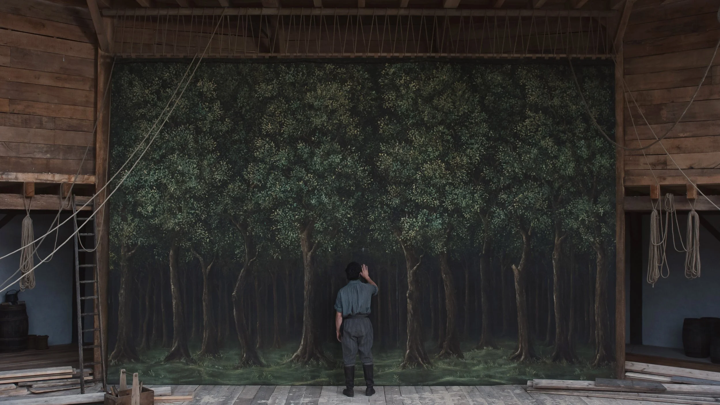 A man stands on a wooden stage facing a large painted forest backdrop, with rows of trees filling the scene and stage ropes and beams visible above, emphasizing the contrast between constructed theater space and imagined nature.