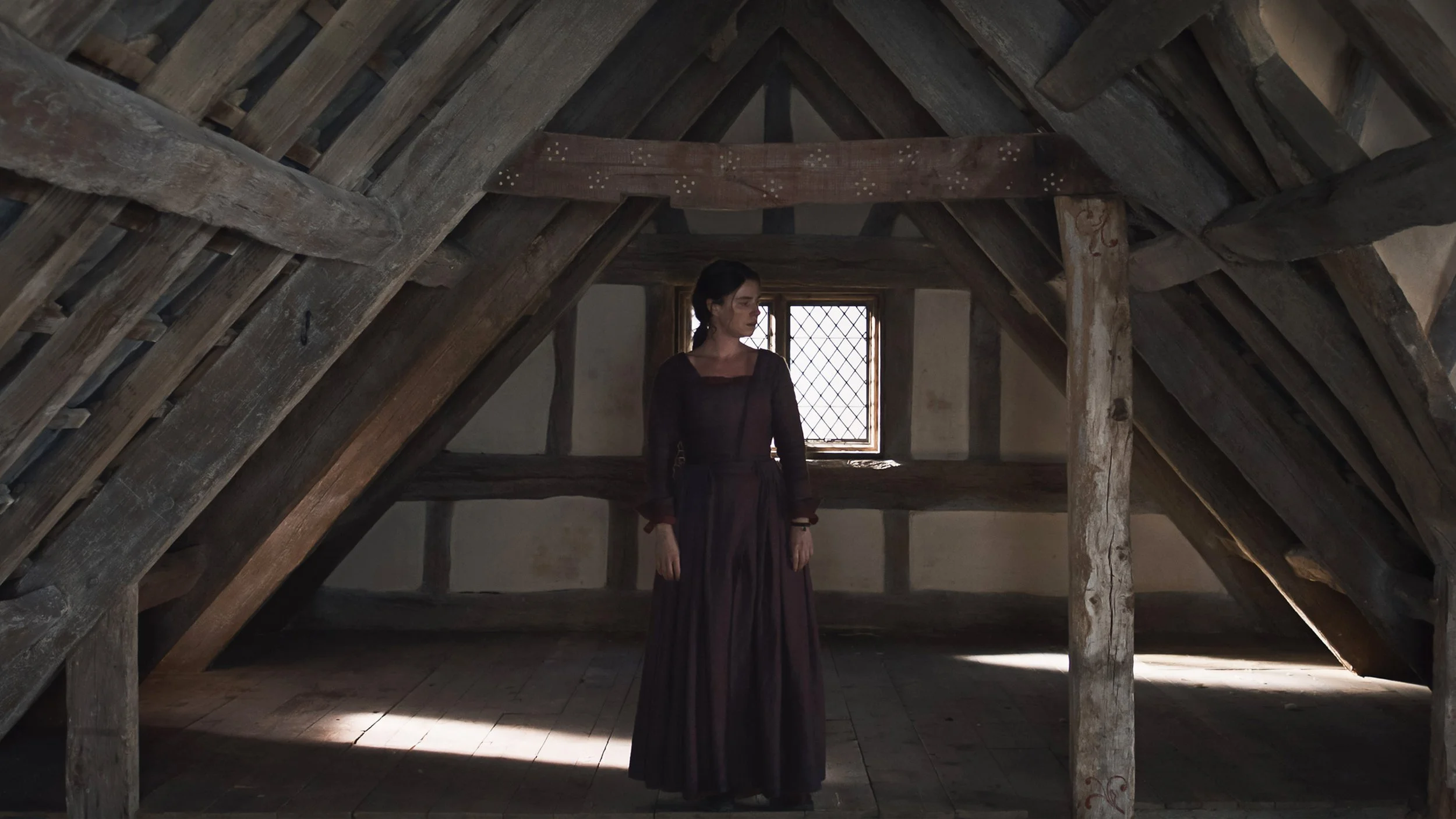 A woman in a dark dress stands alone in a dim wooden attic, framed by heavy timber beams and soft natural light filtering through a small window, creating a quiet and contemplative atmosphere.