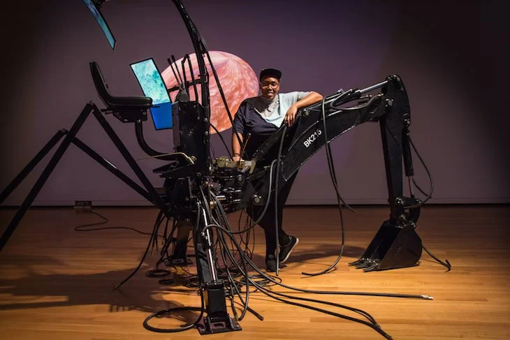 A woman standing behind a technical installation that features screens, wires, stands, and a construction arm.