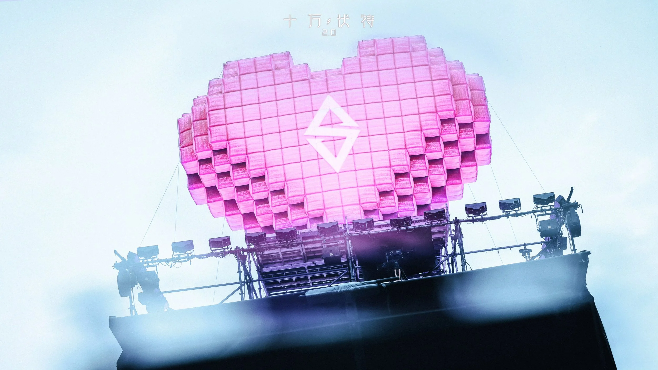 Large pixelated pink heart-shaped stage structure with a lightning-bolt logo at its center, mounted above a scaffolding rig with stage lights, photographed from below against a pale sky.