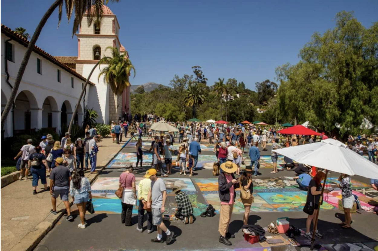 Wide view of outdoor street painting festival with multiple artworks and visitors walking through.