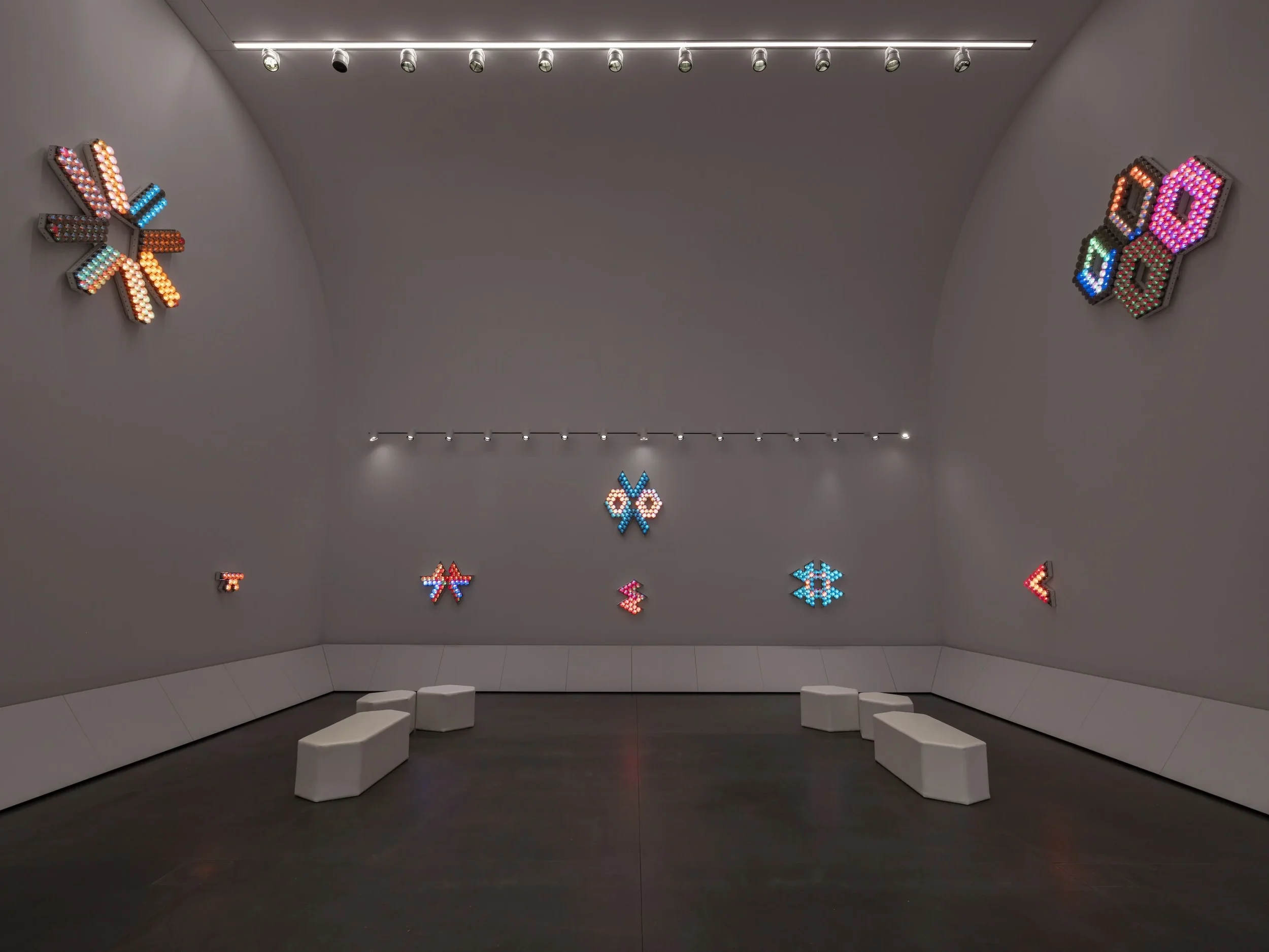 Exhibition space that features large white walls and and geometric seating areas. On the walls hang colorful light sculptures.