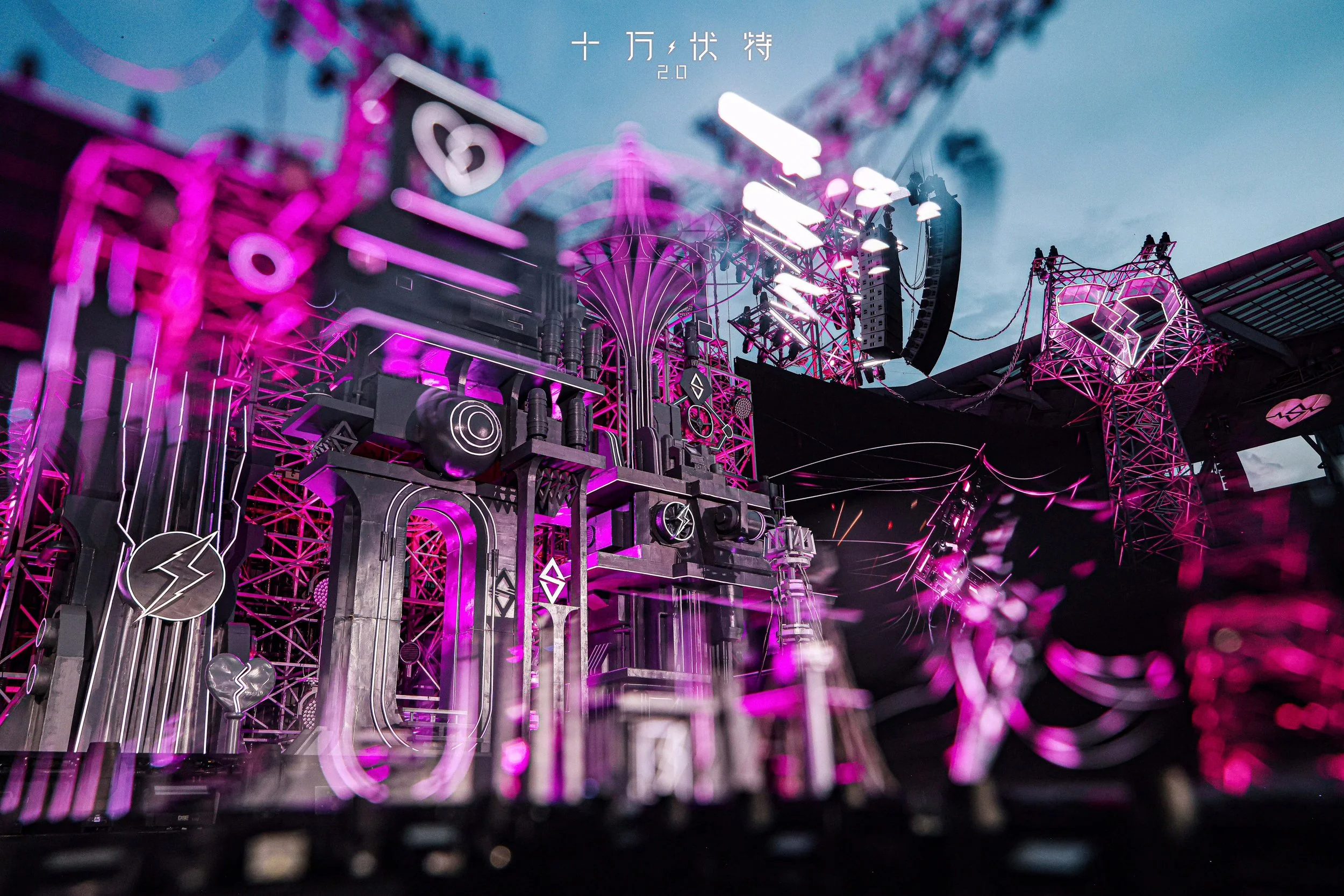 Concert stage design in magenta and purple lighting, with metallic scaffolding, lightning-bolt and broken-heart motifs, and a castle-like tower structure. Chinese text '十万伏特 2.0' appears at top against a dusk sky.