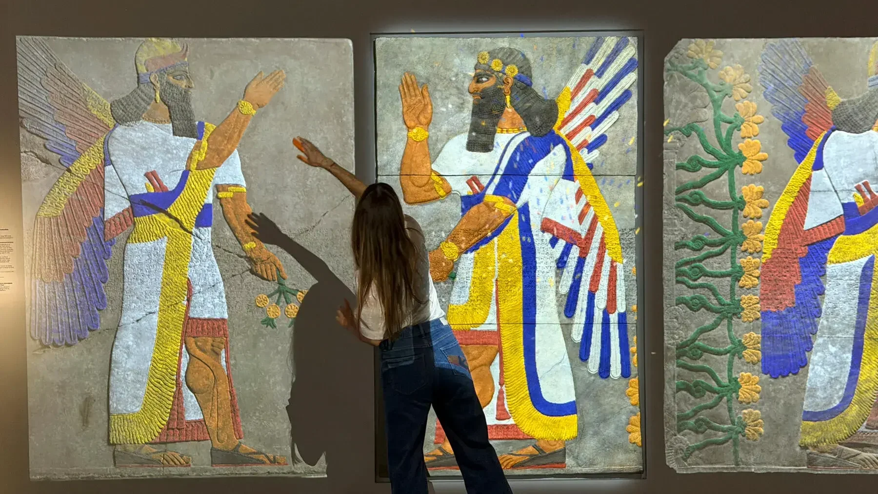 A person waves their hand over a facsimile Assyrian relief depicting a winged figure with vibrant colors.