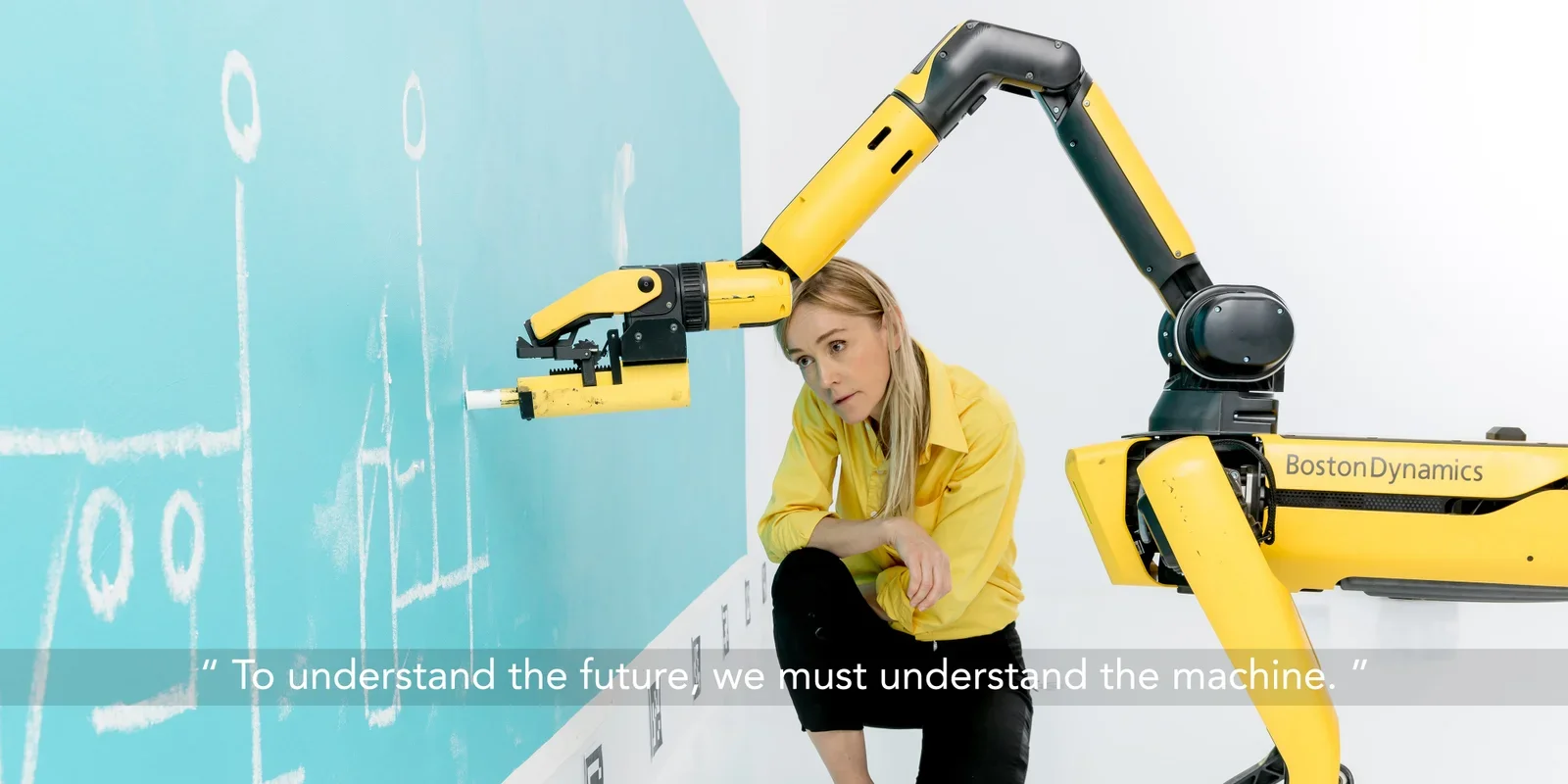 Artist Agnieszka Pilat crouches beside a yellow industrial robotic arm as it draws chalk-like marks on a blue wall, illustrating a collaborative art-making process between a human artist and an AI-controlled robot in a contemporary gallery setting.