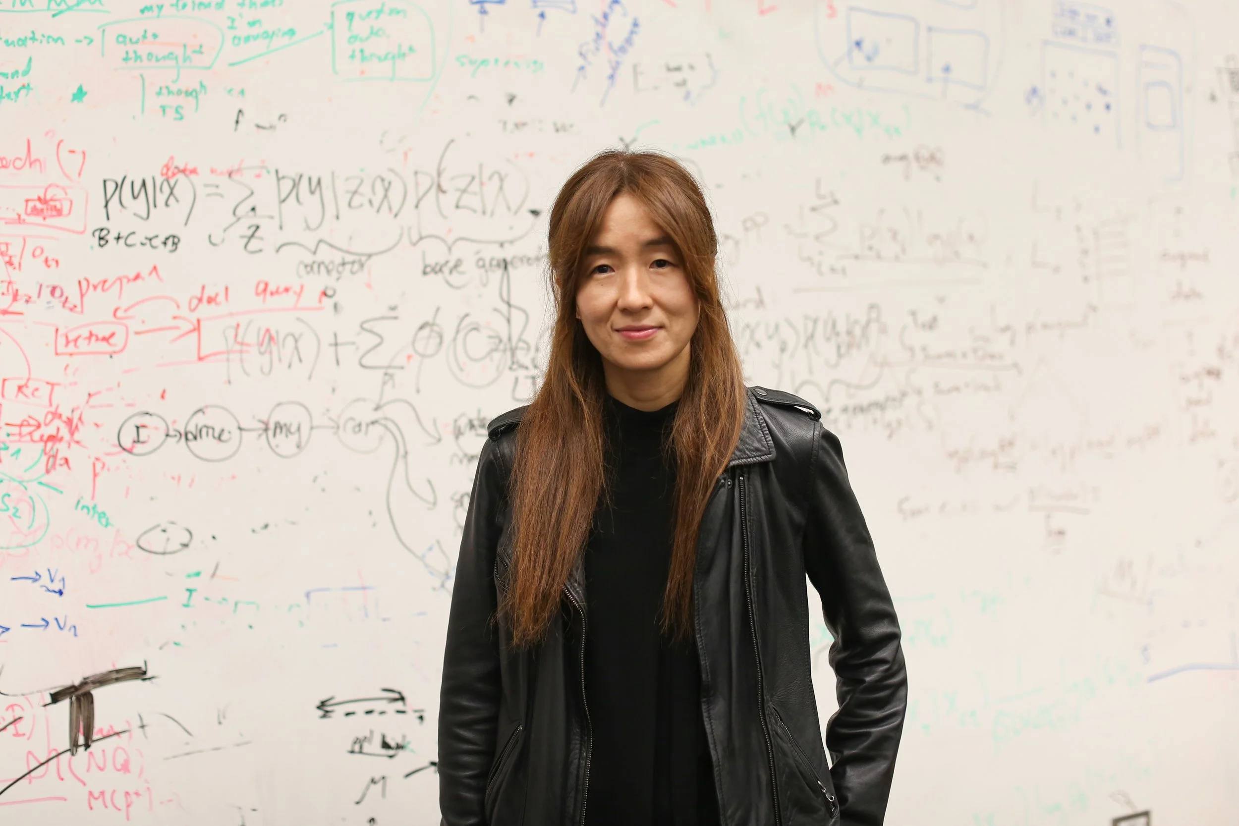 Woman wearing a leather jacket and black shirt, standing in front of a whiteboard with equations