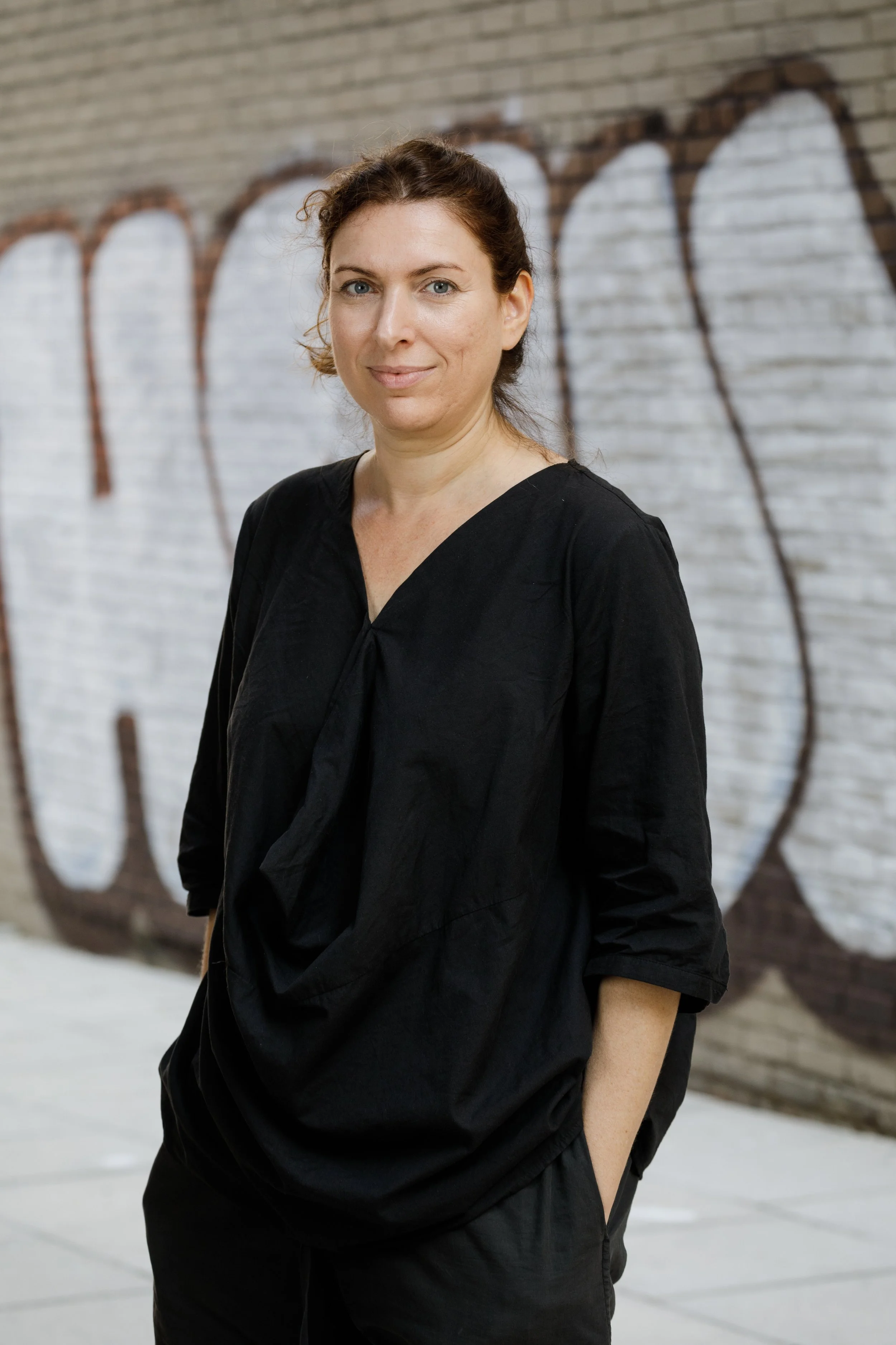 Woman wearing black outfit standing in front off a wall tagged with text.