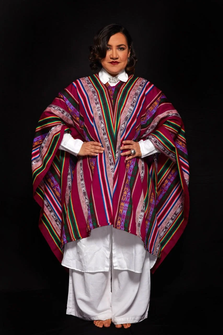 Woman with a colorful garment and white pants, in front of a black background.
