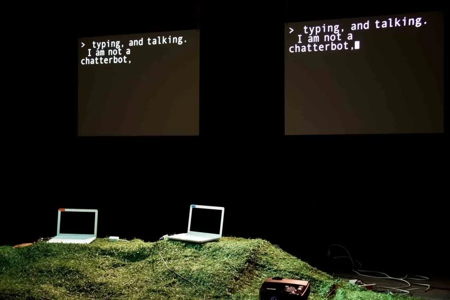 Two laptop computers sitting on grass mound,in front of a black chat screen that says "typing, and talking: I am not a chatterbot,"