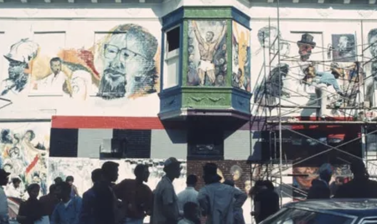Crowd gathered on a city street watching artists paint large-scale murals of Black figures on a building façade, with scaffolding visible and artwork in progress.